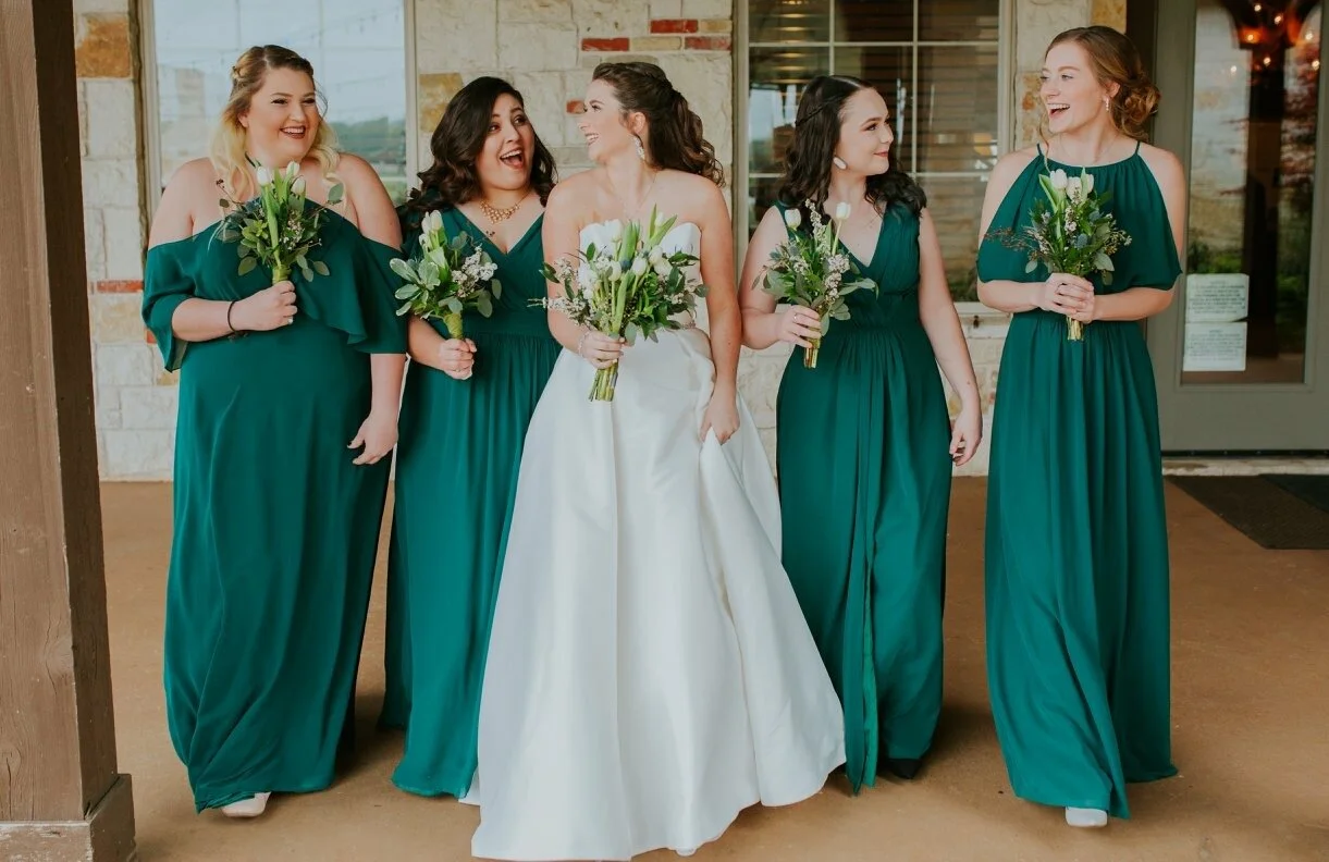 Bride in a classic white wedding dress walks alongside four bridesmaids wearing emerald green gowns and carrying greenery bouquets, capturing a joyful bridal party moment outside the wedding venue.