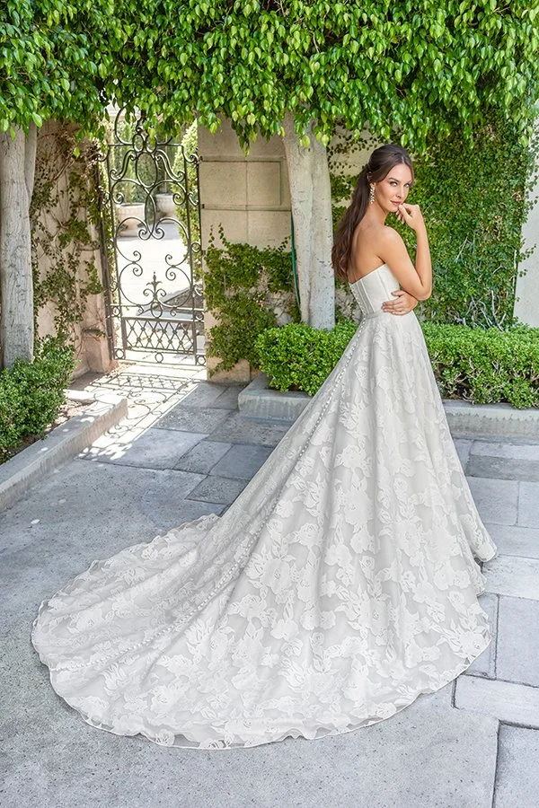 Back view of a bride in a strapless floral jacquard ball gown wedding dress with a structured corset bodice and sweeping cathedral train, highlighting timeless elegance and romantic bridal style in a garden estate setting.