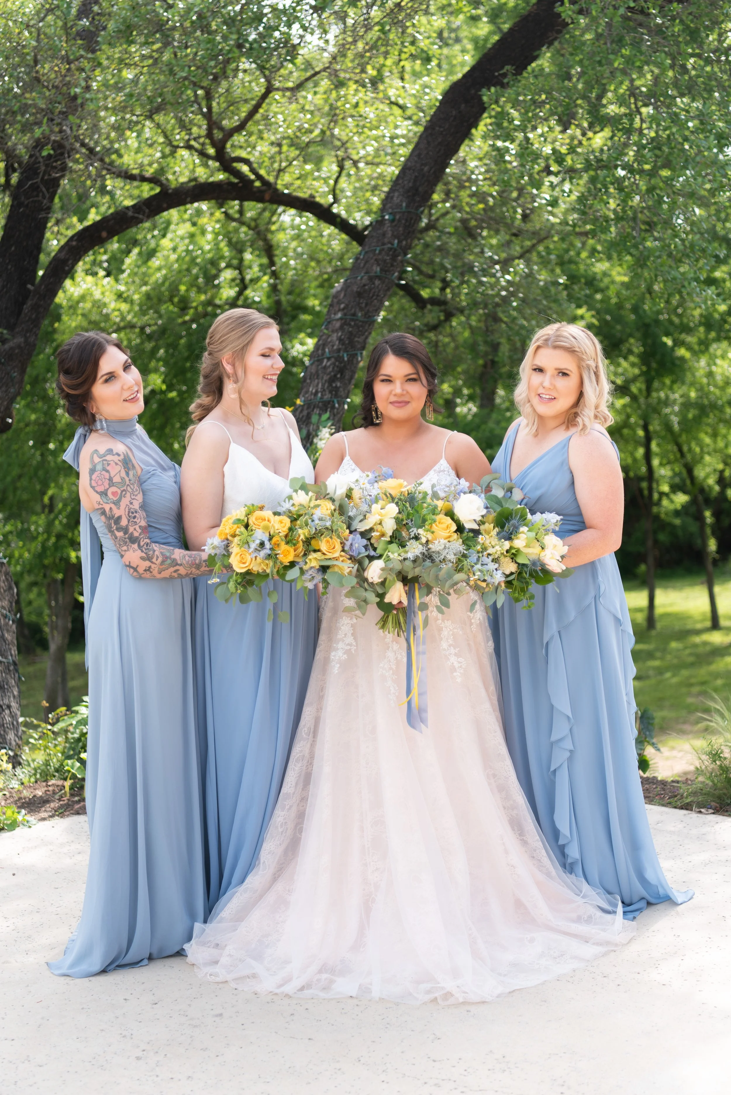 Bride and three bridesmaids pose outdoors in coordinated light blue dresses, showcasing vibrant bouquets of yellow, white, and blue blooms against a lush garden backdrop for a timeless bridal party portrait.