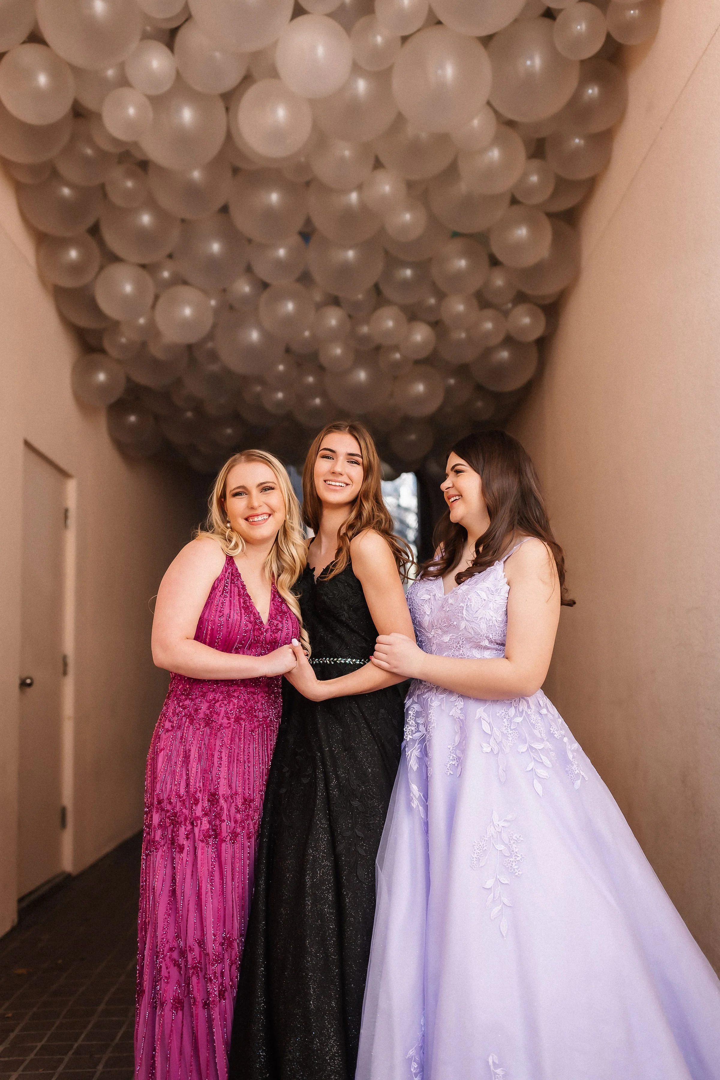 Three friends smile and pose beneath a dramatic ceiling of white balloons, showcasing vibrant formalwear including a pink beaded gown, a black shimmer dress, and a lavender floral ballgown. A stylish prom and special occasion portrait.