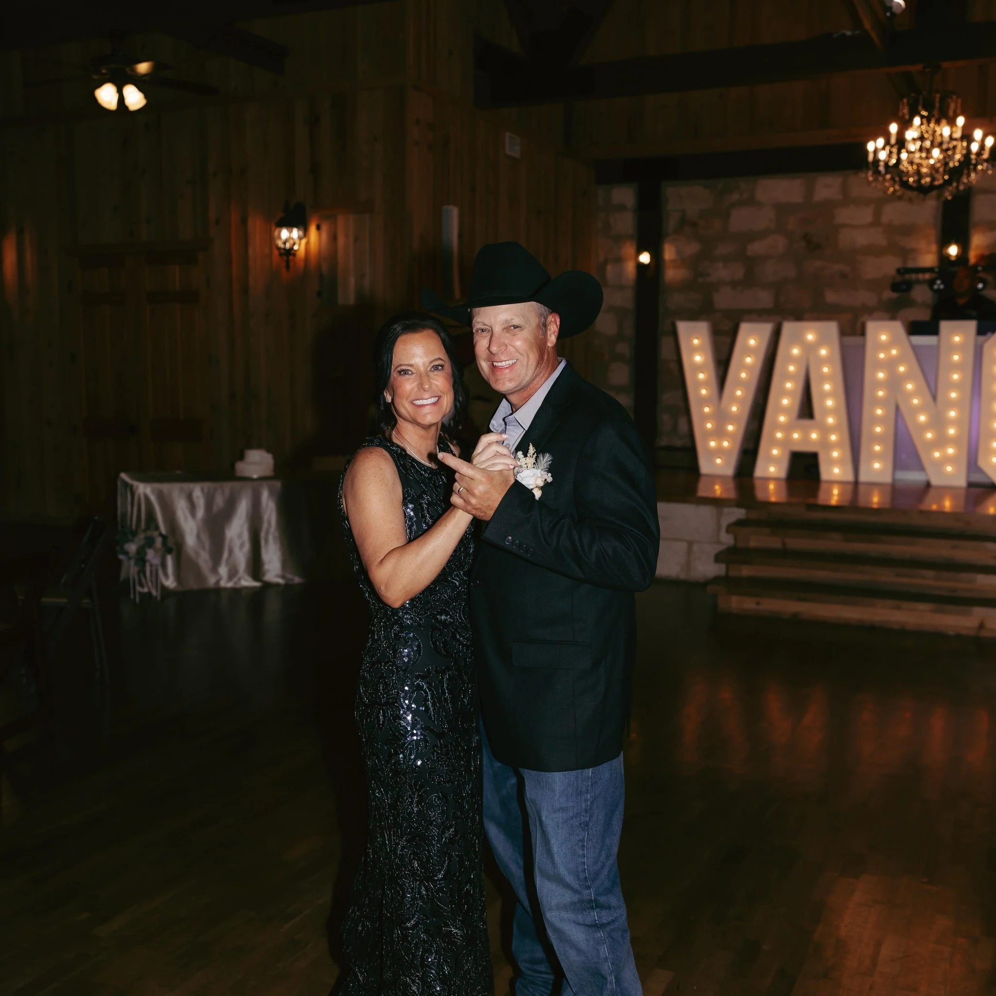 Mother and father of the groom dances together at a wedding reception, the woman wearing a black sequined gown and the man in a cowboy hat, blazer, and jeans. Warm lights and large illuminated letters in the background.