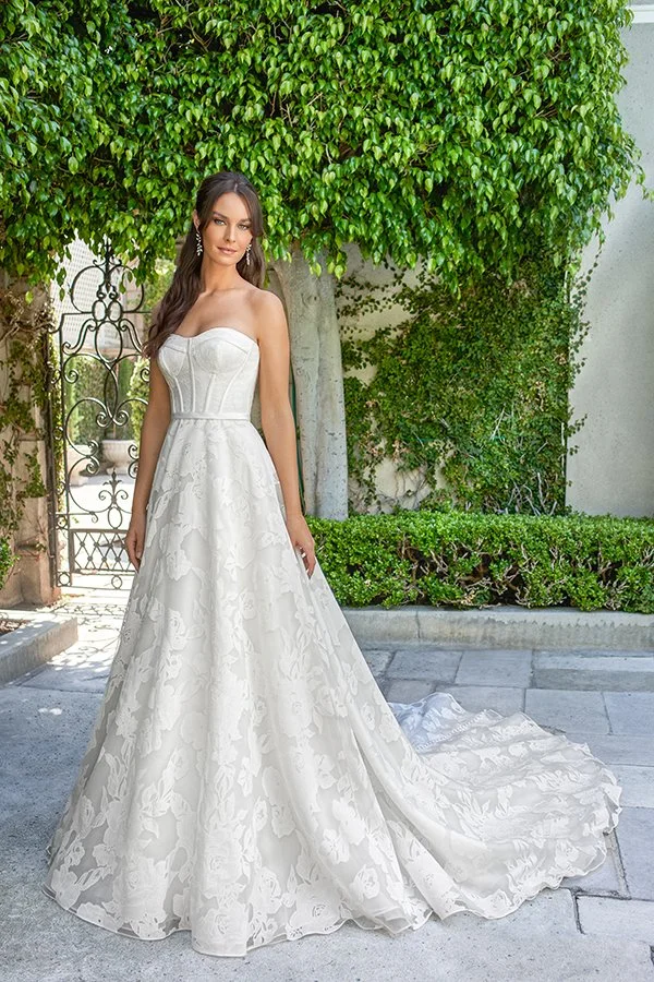 Bride models a strapless floral jacquard ball gown wedding dress with a structured corset bodice, sweetheart neckline, and flowing train in a beautiful garden courtyard, showcasing timeless elegance and romantic bridal style.