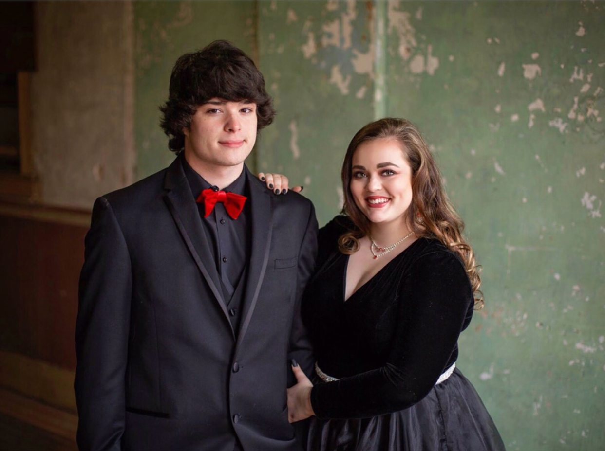 Prom couple posing in coordinated black formalwear, featuring a sleek black suit accented with a bold red bow tie and a black velvet gown with a flowing tulle skirt, creating a timeless and elegant formal portrait.