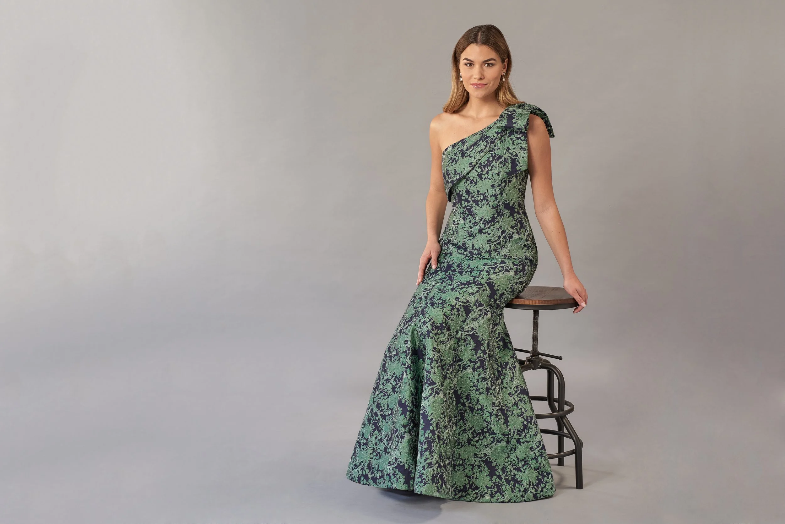 Model wearing a sophisticated one-shoulder mermaid gown with a green and black floral brocade pattern, showcasing an elegant fitted silhouette ideal for mother-of-the-bride and formal event styling.