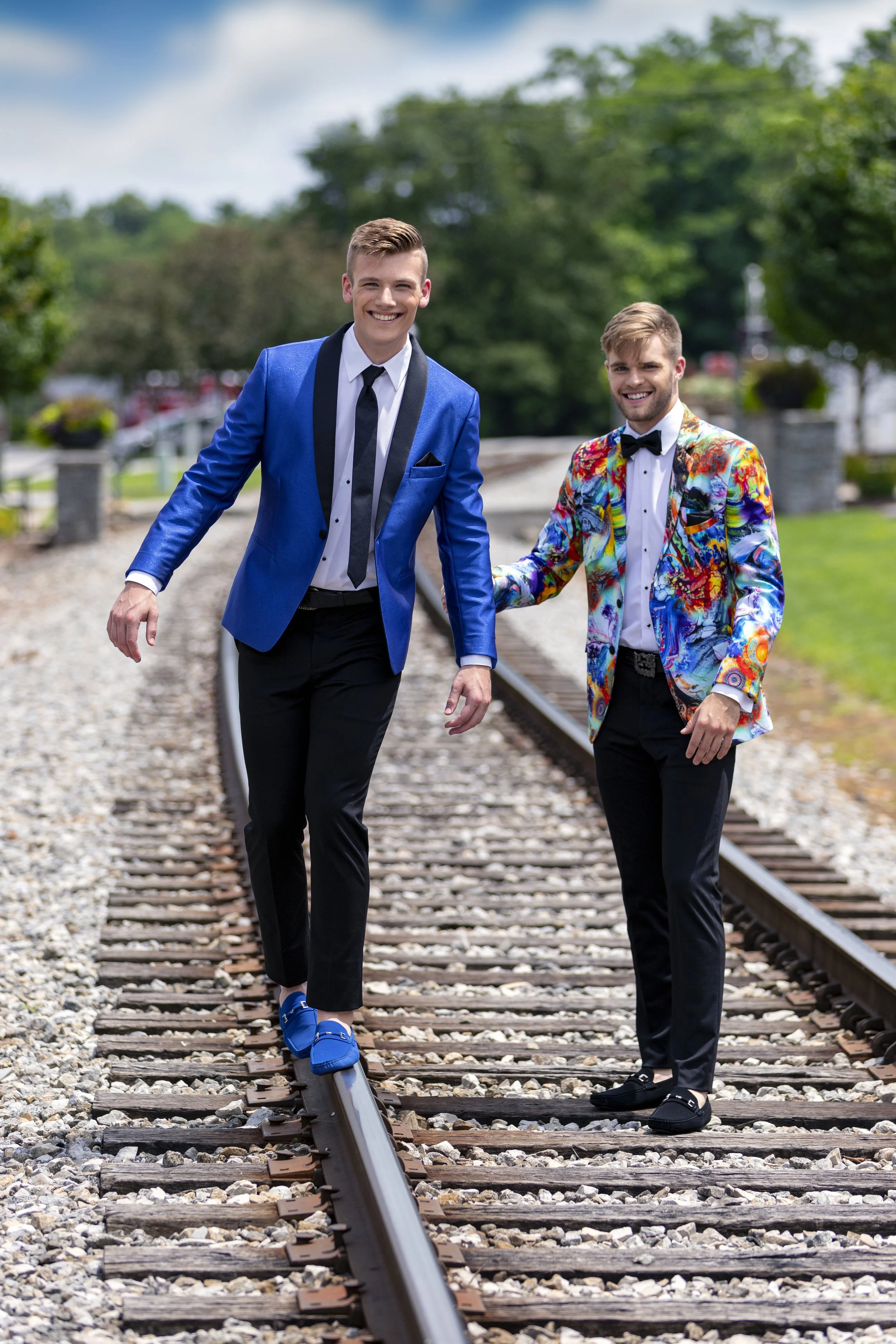 Two young men showcase contrasting prom styles on railroad tracks, featuring a classic royal blue tuxedo jacket and a vibrant abstract-print tuxedo jacket, highlighting modern and fashion-forward formalwear.