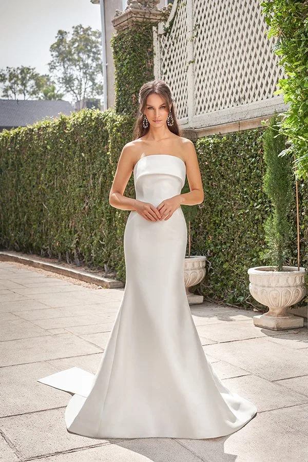 Bride models a strapless satin fit-and-flare wedding dress with a modern folded neckline and sweeping train in a sophisticated garden estate, showcasing timeless bridal style, minimalist elegance, and luxury wedding dress inspiration.