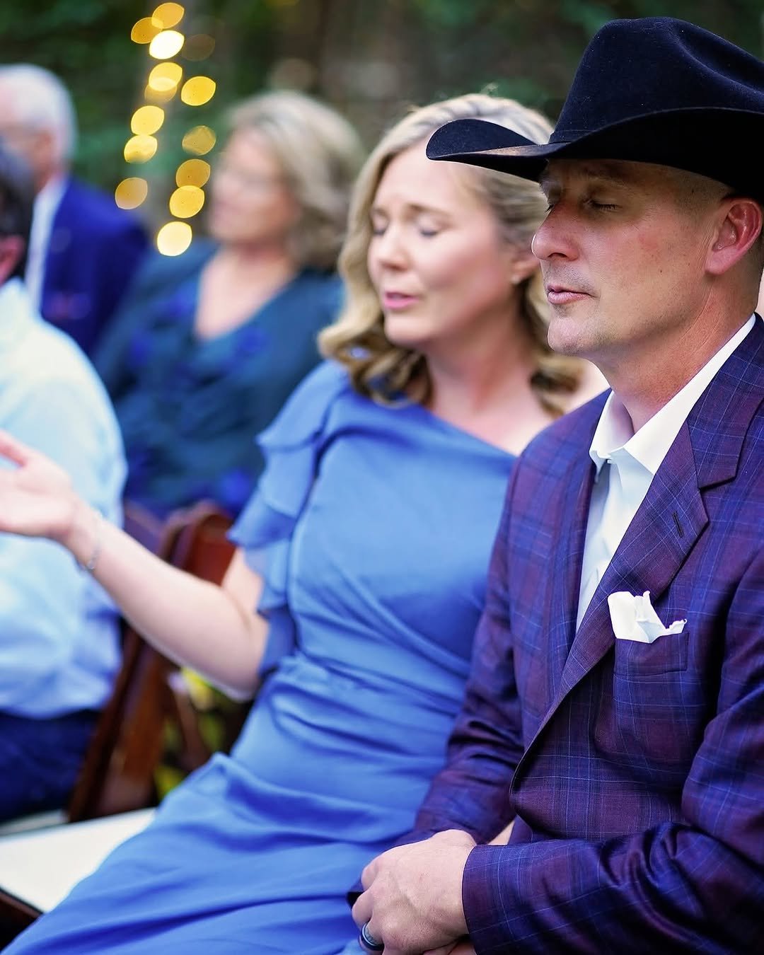 A couple sits together with eyes closed during a wedding ceremony, the woman in a blue dress with hands lifted in prayer and the man in a cowboy hat and plaid suit, soft lights glowing in the background.