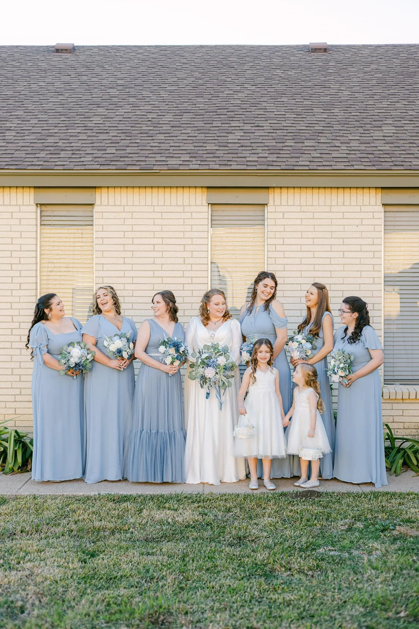 Bride poses with her bridal party featuring bridesmaids in elegant dusty blue gowns and two flower girls in white dresses, holding coordinated blue and white bouquets for a charming outdoor wedding portrait.