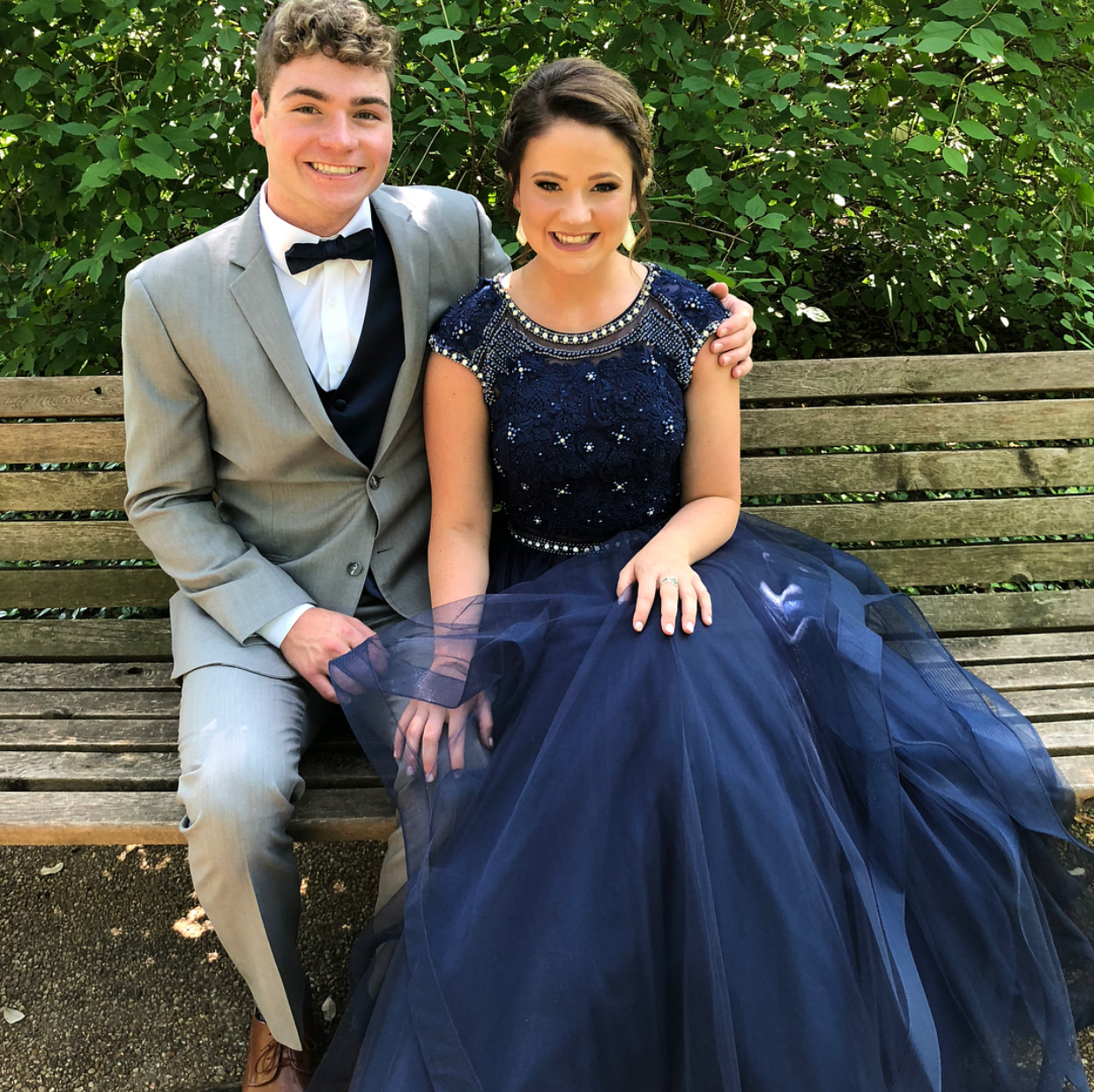 A smiling young couple poses on a wooden bench. The young woman wears an elegant navy blue gown with intricate beading and a flowing tulle skirt, while her date wears a classic light gray suit and black bow tie, creating a timeless prom portrait.