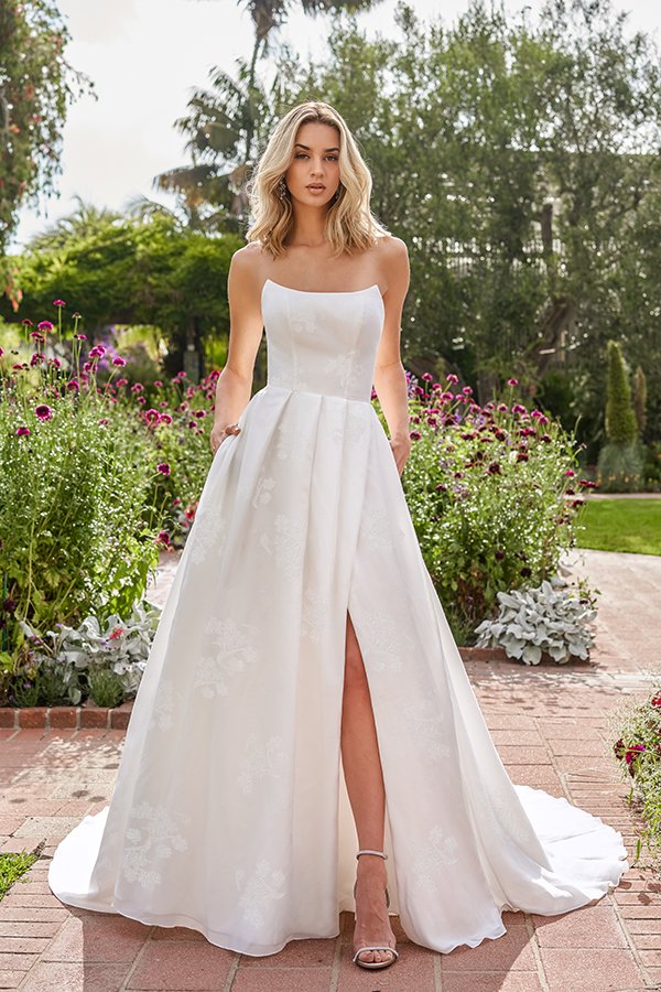 Bride modeling a modern strapless A-line wedding dress with a clean straight neckline, subtle floral jacquard fabric, hidden pockets, a thigh-high slit, and a flowing chapel train, photographed in a beautiful flower-filled garden.