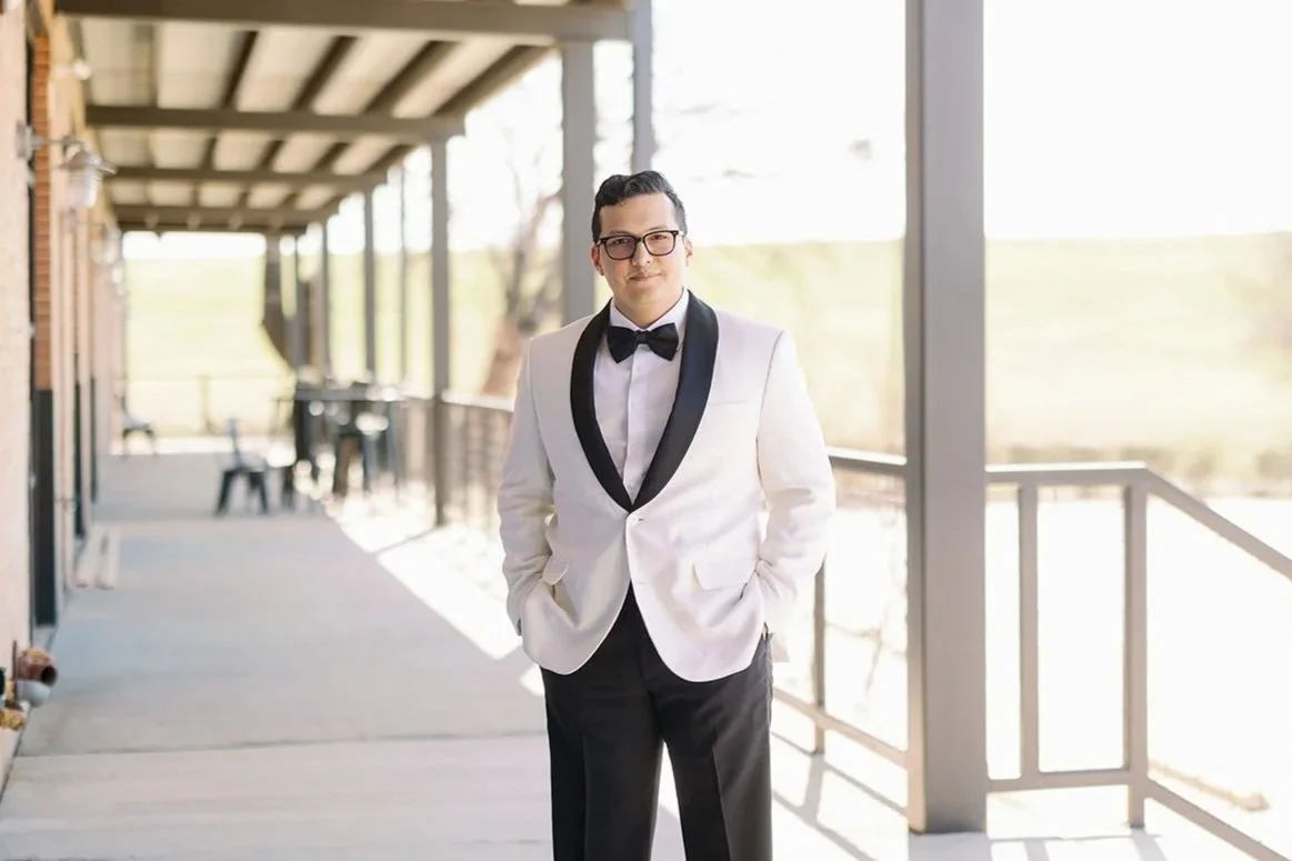 Groom showcasing a sophisticated white tuxedo jacket with black satin lapels, paired with a black bow tie and trousers, creating a timeless groom look on an elegant outdoor venue walkway.