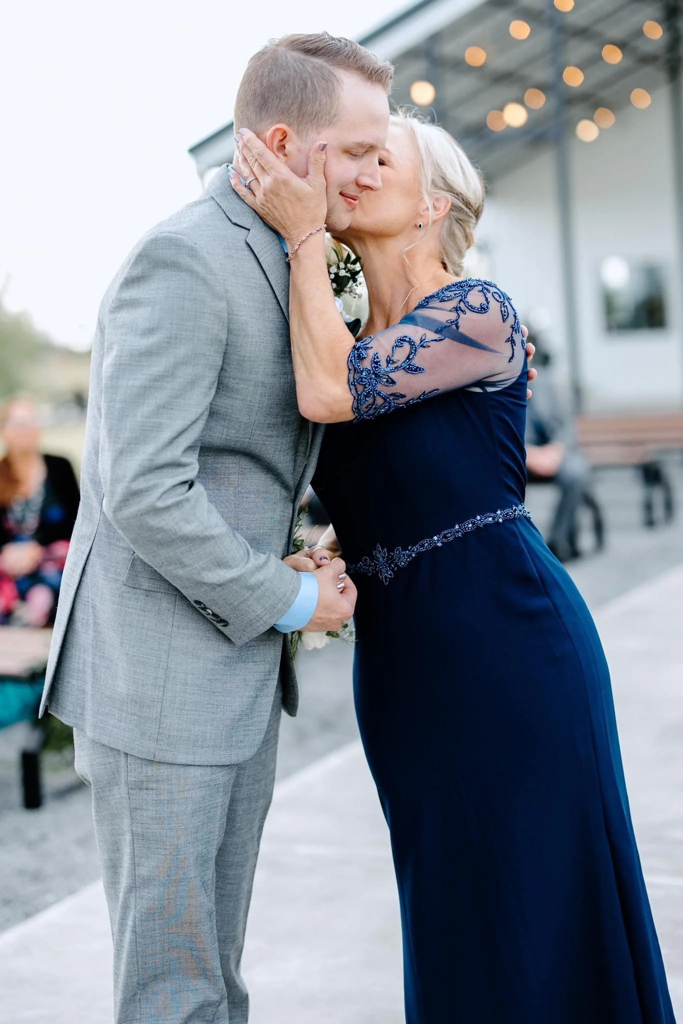 A mother in a navy beaded gown kisses her son’s cheek during an outdoor wedding moment. He wears a light gray suit and holds her hands as they share an emotional embrace with soft lights and guests in the background.