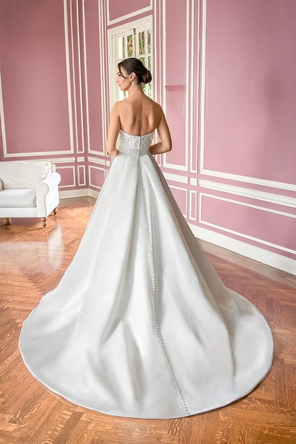 Bride showcasing the back of a strapless satin ball gown wedding dress featuring a floral appliqué bodice, button-back closure, and dramatic cathedral train, photographed in an elegant blush pink bridal suite.