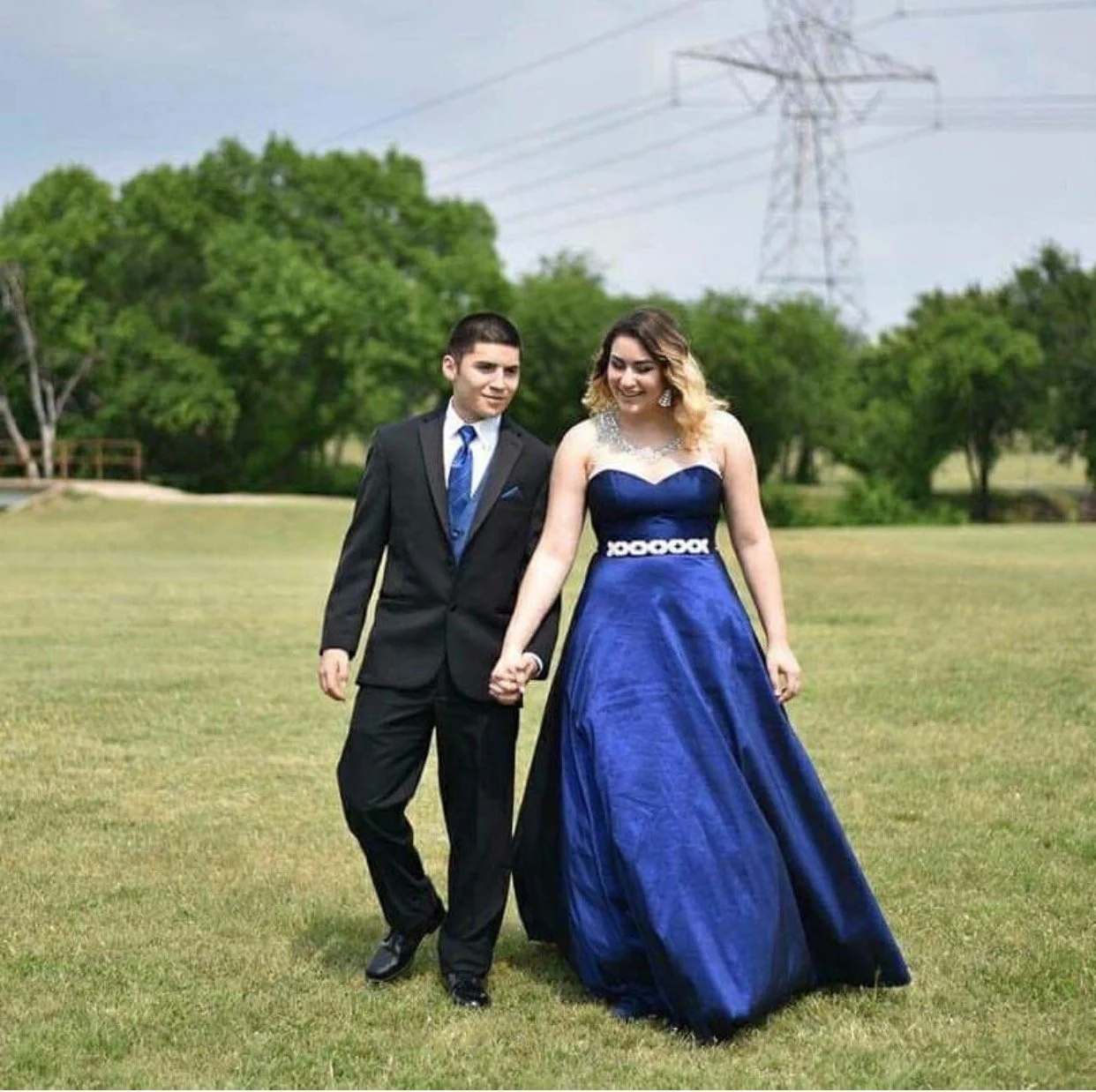 Prom couple strolls hand in hand across an open field, showcasing coordinated formalwear with a royal blue strapless ball gown and a classic black suit accented by a matching blue tie for a timeless prom portrait.