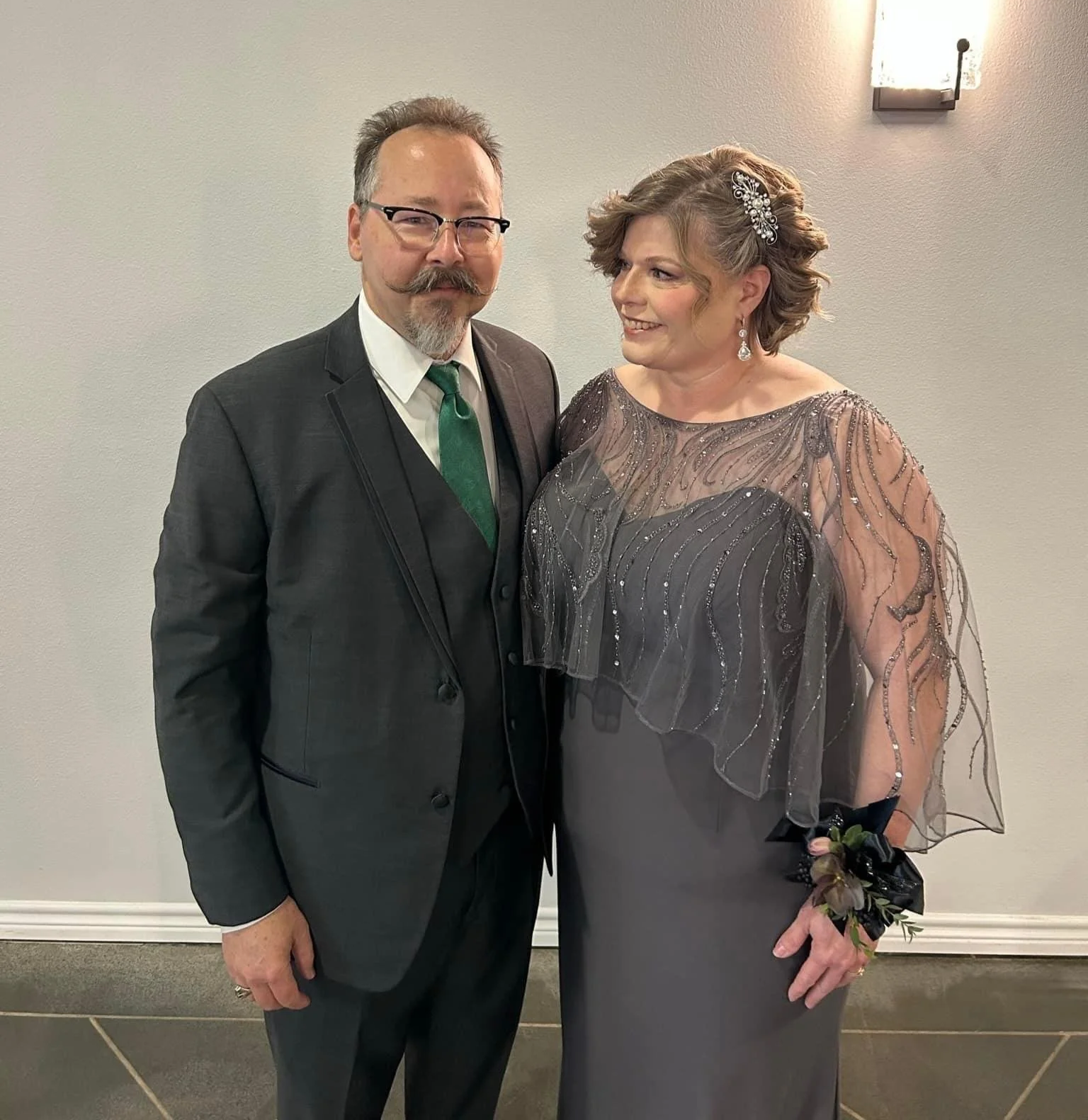 A couple stands together indoors, smiling warmly. The woman wears a charcoal mother-of-the-bride gown with a beaded capelet and floral wrist corsage, while the man wears a dark suit with a green tie. Soft wall lighting creates an elegant backdrop.