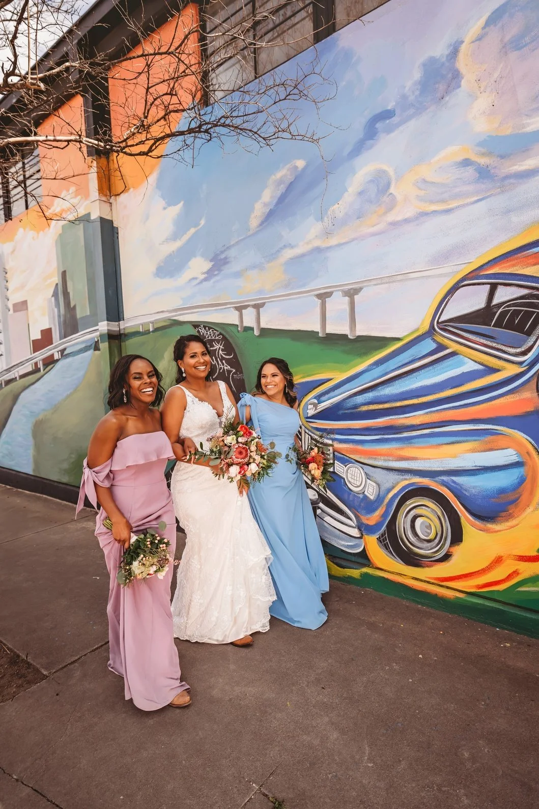 Bride and her bridesmaids stroll together with floral bouquets in front of a vibrant mural, creating a fun and colorful urban wedding portrait featuring coordinated bridal party fashion.