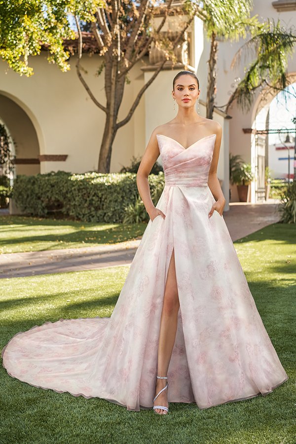 Bride modeling a romantic blush floral A-line wedding dress with a strapless sweetheart neckline, softly draped bodice, hidden pockets, a thigh-high slit, and a flowing chapel train in a beautiful outdoor garden courtyard.
