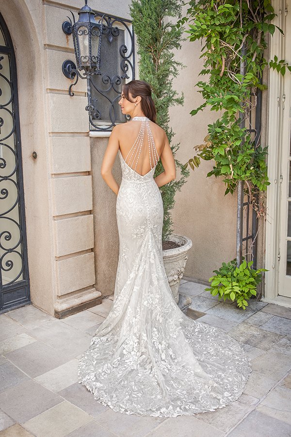 Back view of a bride in a lace halter mermaid wedding dress featuring a dramatic open back with cascading pearl straps and a lace train, showcasing elegant bridal style at a sophisticated garden estate.