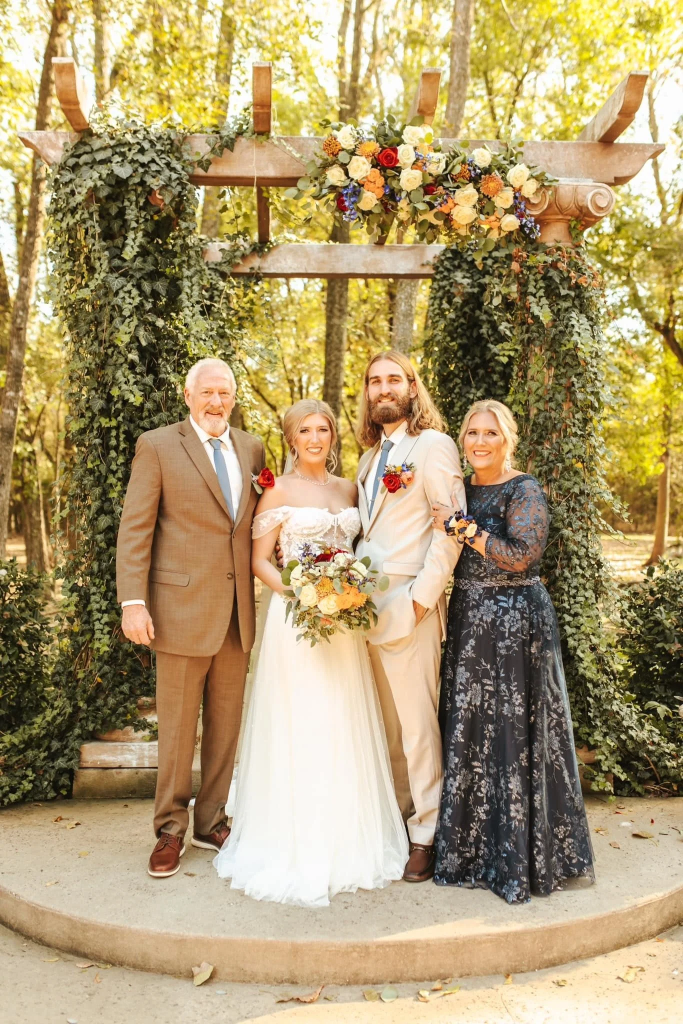 The bride and groom pose with their parents under a floral-decorated wooden arbor, surrounded by greenery. The bride holds a bouquet of warm-toned flowers as everyone smiles at the camera.