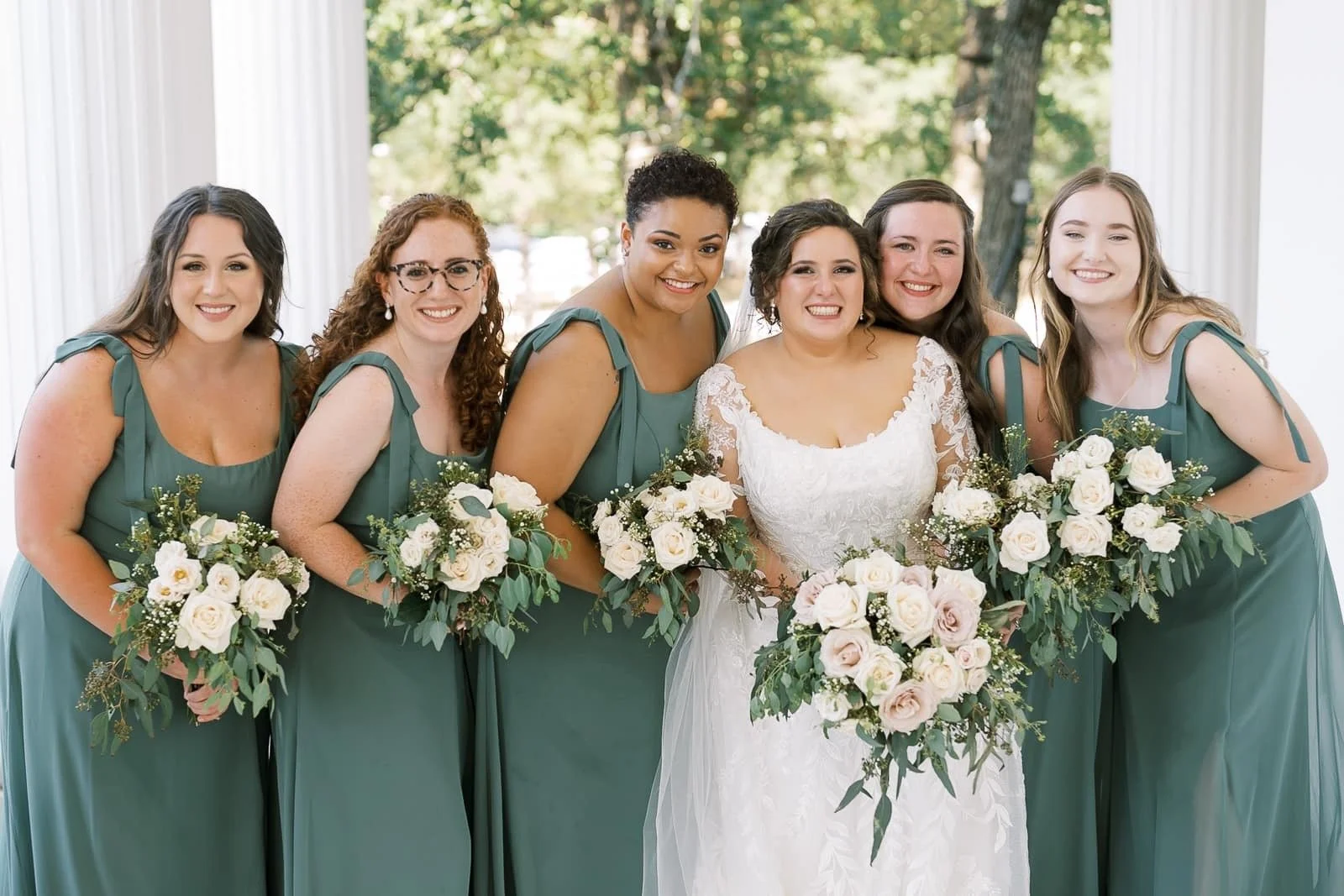 Bride in an elegant lace wedding gown stands with five bridesmaids in coordinated sage green dresses, holding lush white rose bouquets for a timeless bridal party portrait at an outdoor wedding venue.