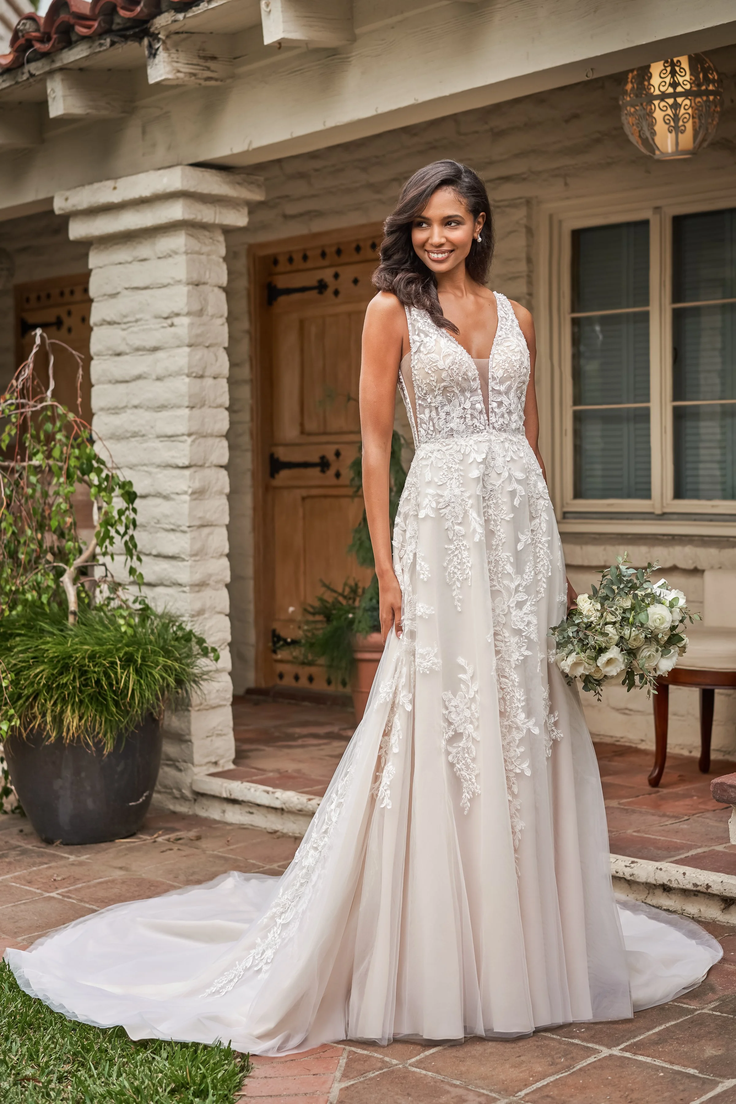 Bride poses outside a rustic stone wedding venue in a floral lace A-line wedding dress with a deep V-neckline and flowing train, holding a white bouquet to showcase timeless bridal fashion and elegant outdoor wedding inspiration.