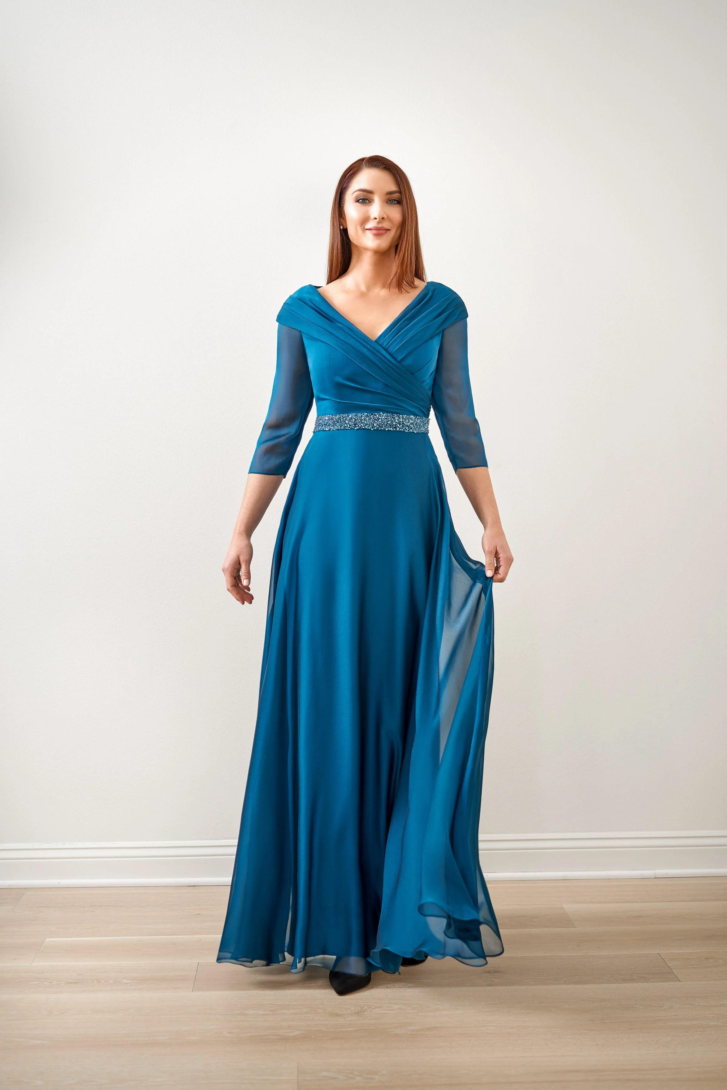 Front view of a teal mother of the bride gown with an off-the-shoulder crossover bodice, sheer three-quarter sleeves, sparkling beaded waistband, and flowing chiffon A-line skirt.