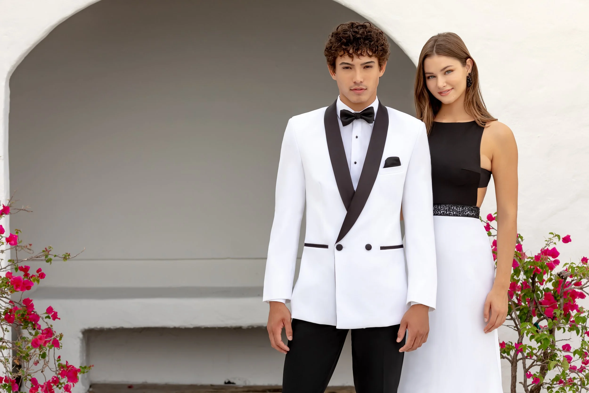 Prom couple posing in sophisticated black-and-white formalwear, featuring a white double-breasted tuxedo with black shawl lapels and a sleek color-block evening gown, set against a bright architectural backdrop with vibrant flowers.