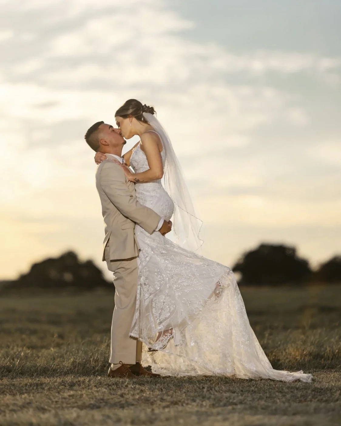 Groom in a tan suit lifts his bride in a lace wedding gown and flowing veil for a romantic sunset kiss, creating a timeless outdoor wedding portrait filled with golden-hour warmth.