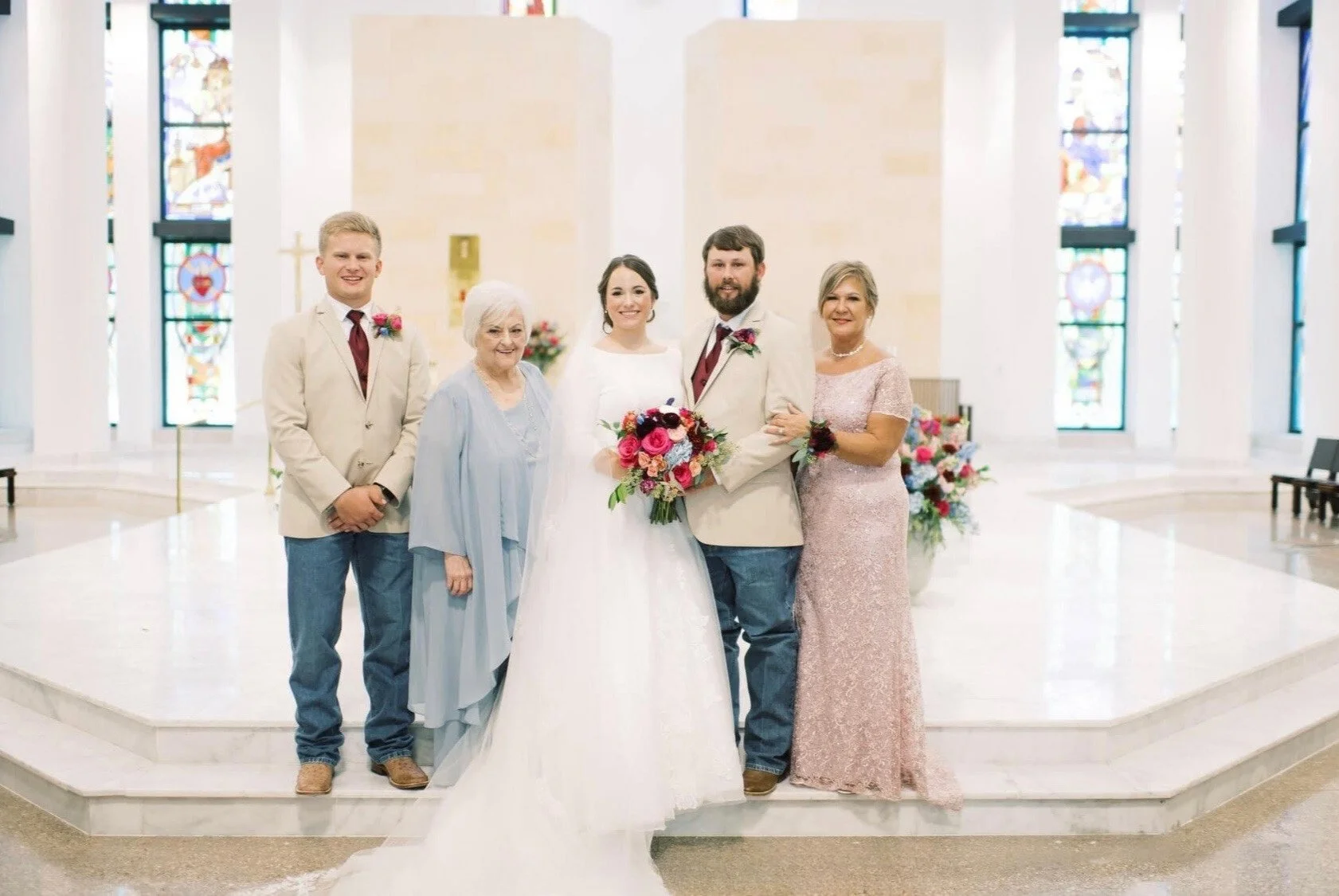 Bride and groom pose with family members in a white, stained-glass church. The mother of the groom wears a blush lace gown, and another family member wears a soft blue ensemble as they gather for a joyful wedding portrait.