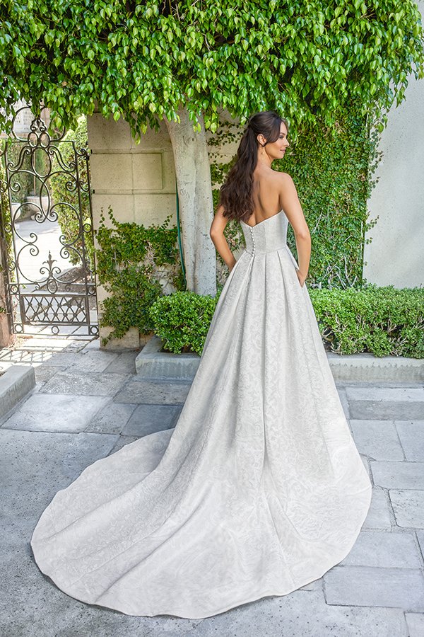 Bride showcasing the back of a strapless jacquard ball gown wedding dress with a structured bodice, hidden pockets, and a sweeping cathedral train in a romantic garden courtyard.