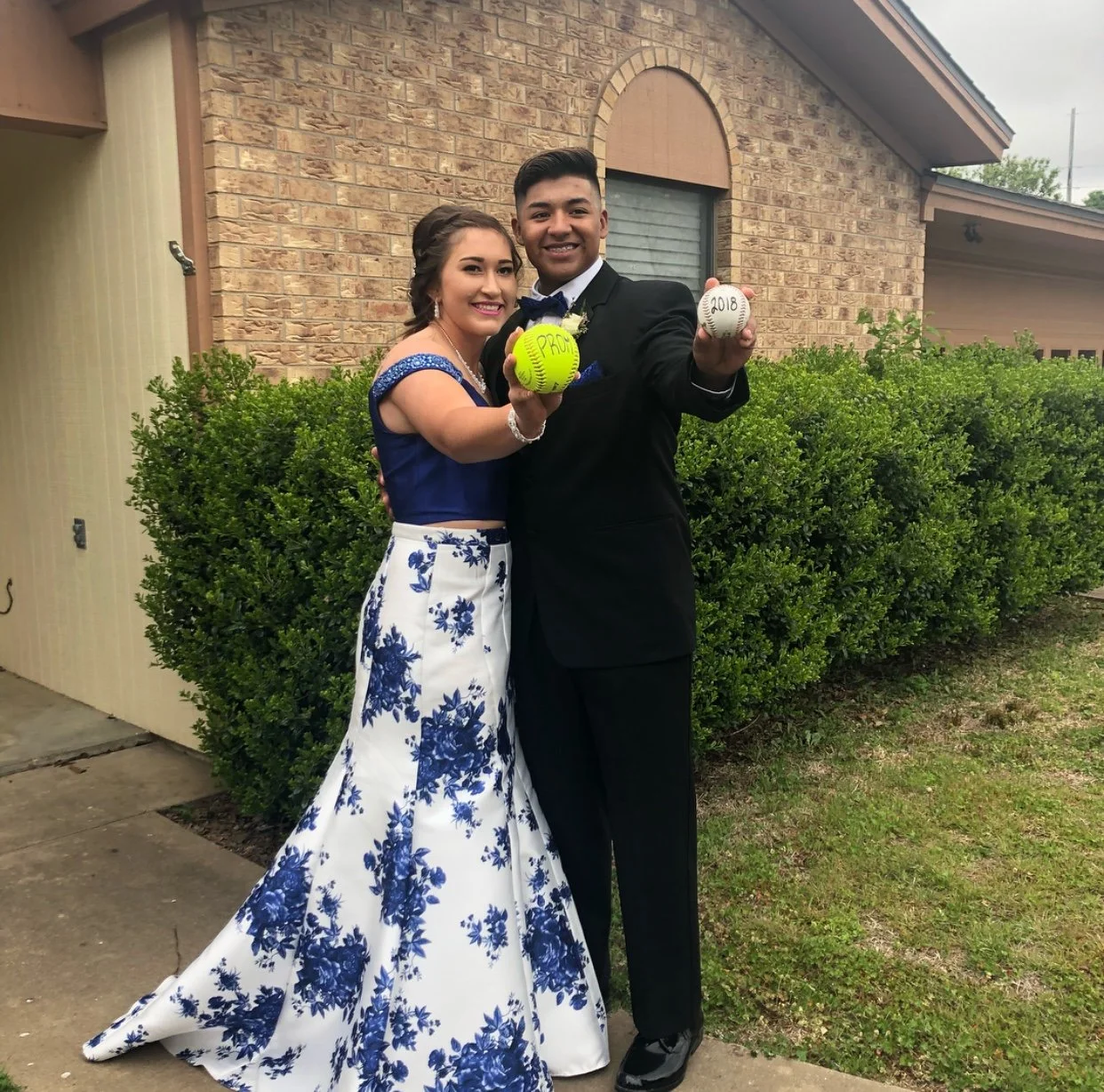 Prom couple showcases a playful sports-inspired pose, wearing a black suit and a blue-and-white floral formal gown while holding a softball and baseball, blending athletic personality with elegant prom fashion.