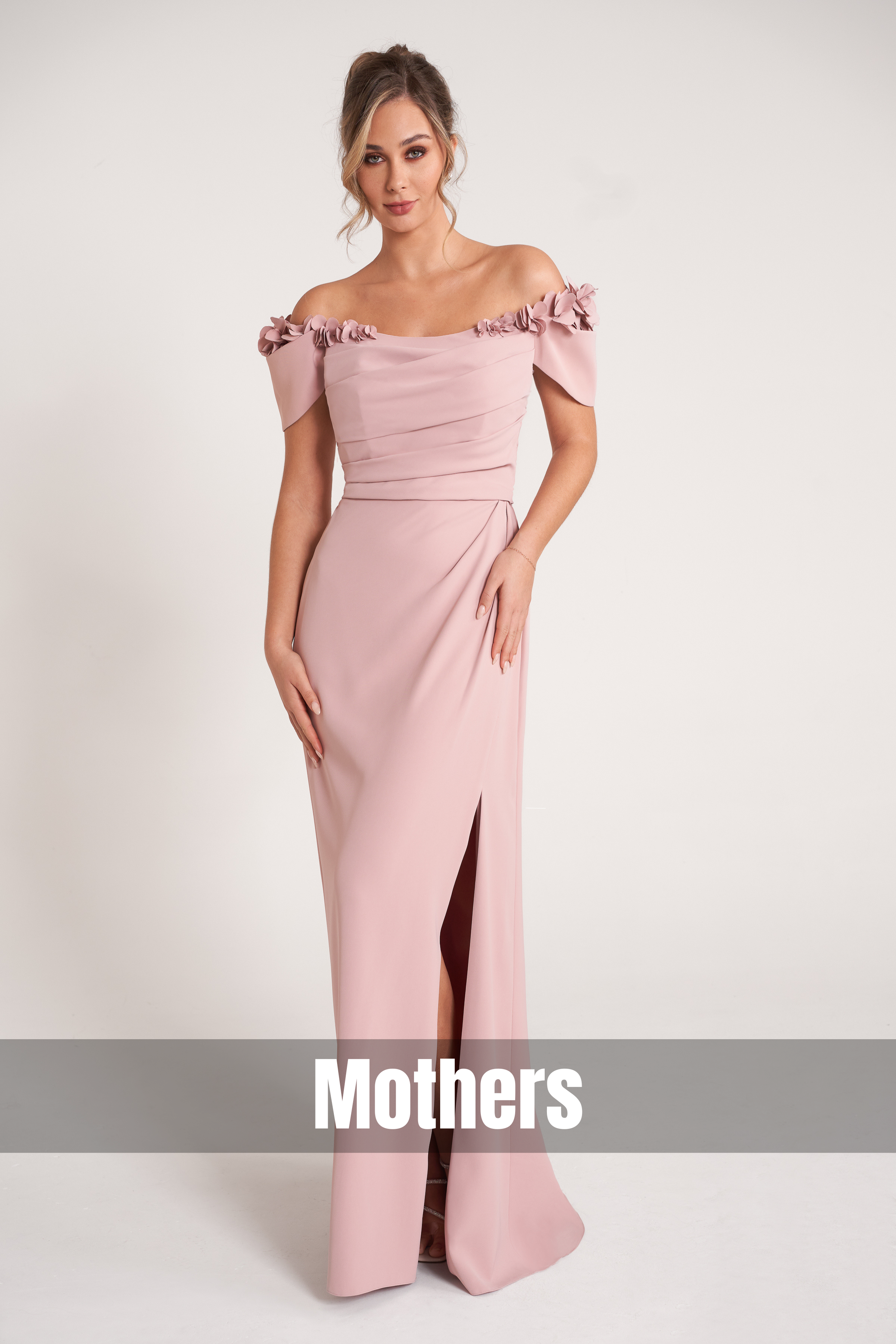 Model wearing a blush pink off-the-shoulder mother-of-the-bride gown featuring floral appliqué details, a softly draped bodice, and a floor-length skirt with a front slit on a neutral studio backdrop.