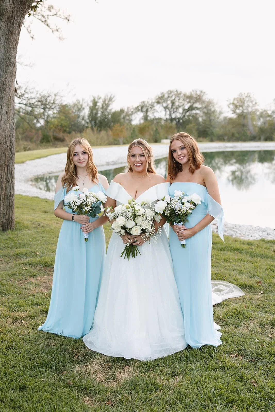 Bride in an elegant off-the-shoulder wedding gown poses with two bridesmaids in soft blue dresses, holding white floral bouquets beside a scenic pond for a classic outdoor wedding portrait.
