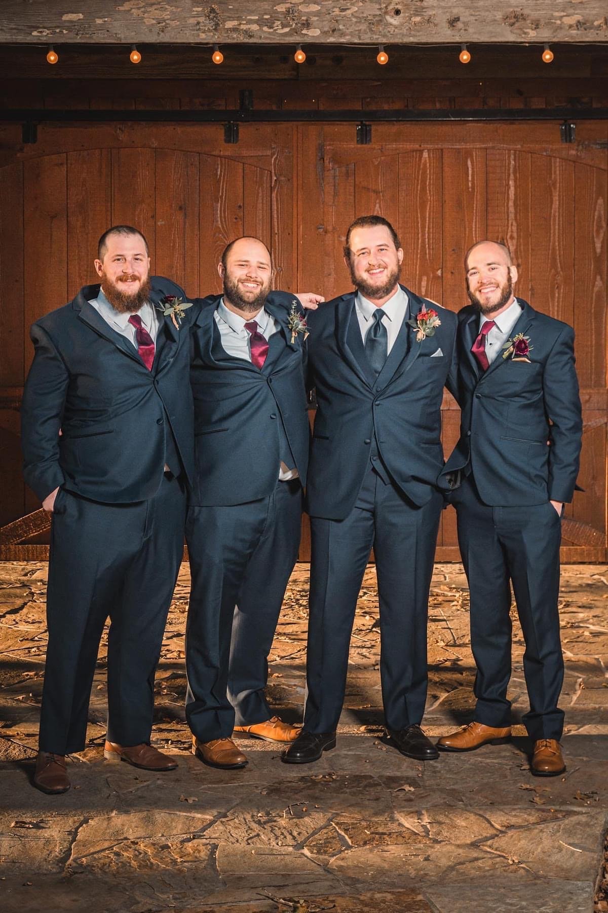 Groom and his groomsmen in coordinated navy blue suits with burgundy ties and matching boutonnieres pose together against a rustic barn backdrop, showcasing classic wedding party style and camaraderie.