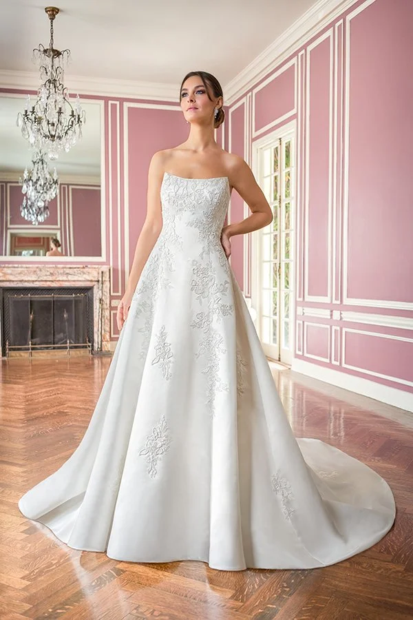 Bride in a square neckline satin A-line wedding dress featuring delicate floral appliqués, a structured bodice, and a flowing chapel train, photographed in a romantic blush pink bridal suite.