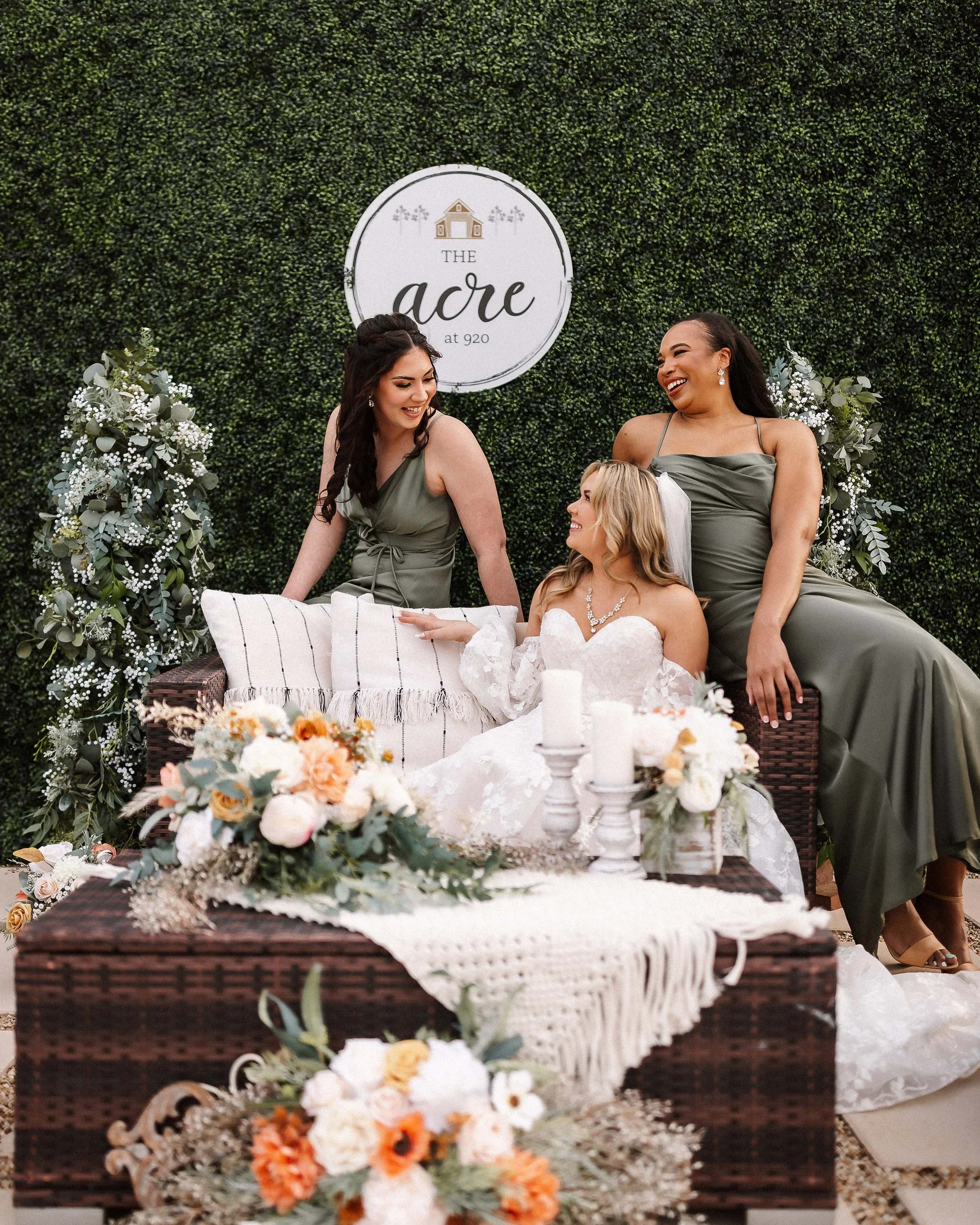 Bride and two bridesmaids share a joyful moment on a beautifully styled wedding lounge featuring sage green dresses, lush florals, candles, and a greenery backdrop, showcasing elegant wedding décor inspiration.