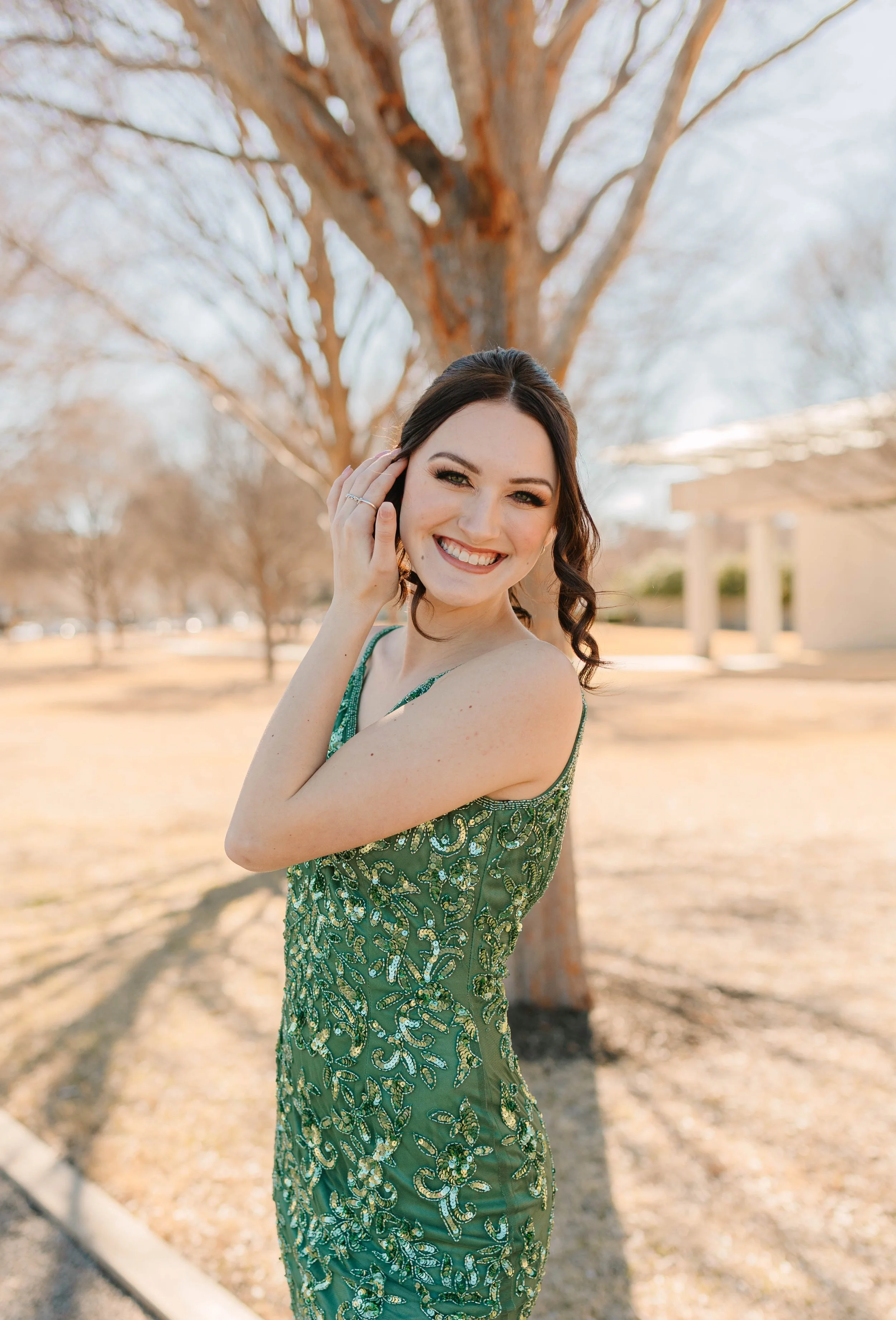 A smiling young woman poses outdoors in a stunning emerald green beaded gown featuring intricate floral embroidery and sparkling embellishments. The sunlit portrait highlights formal fashion, elegance, and special occasion style in a natural setting.