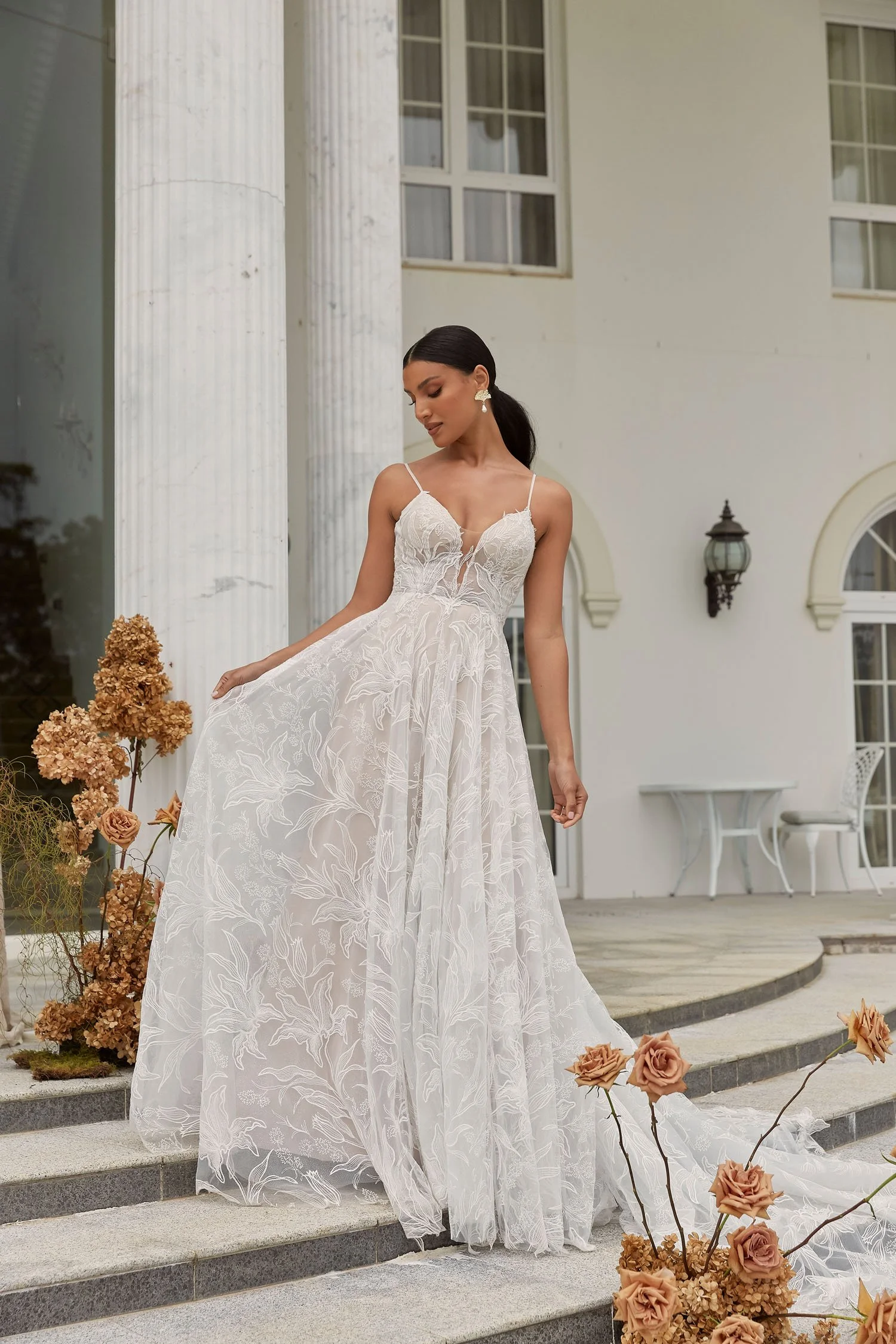Bride models a floral lace A-line wedding dress with delicate spaghetti straps, an illusion bodice, and a flowing train on the steps of a grand estate, showcasing romantic bridal fashion, elegant wedding gowns, and timeless wedding inspiration.