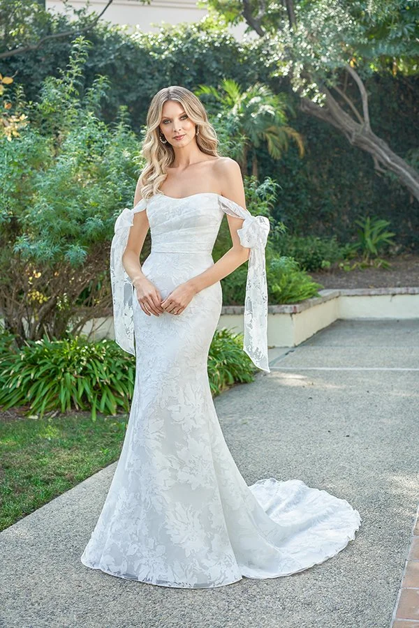 Bride in an off-the-shoulder jacquard fit-and-flare wedding dress featuring detachable bow sleeves, a straight neckline, and a flowing chapel train, photographed in a romantic garden setting.