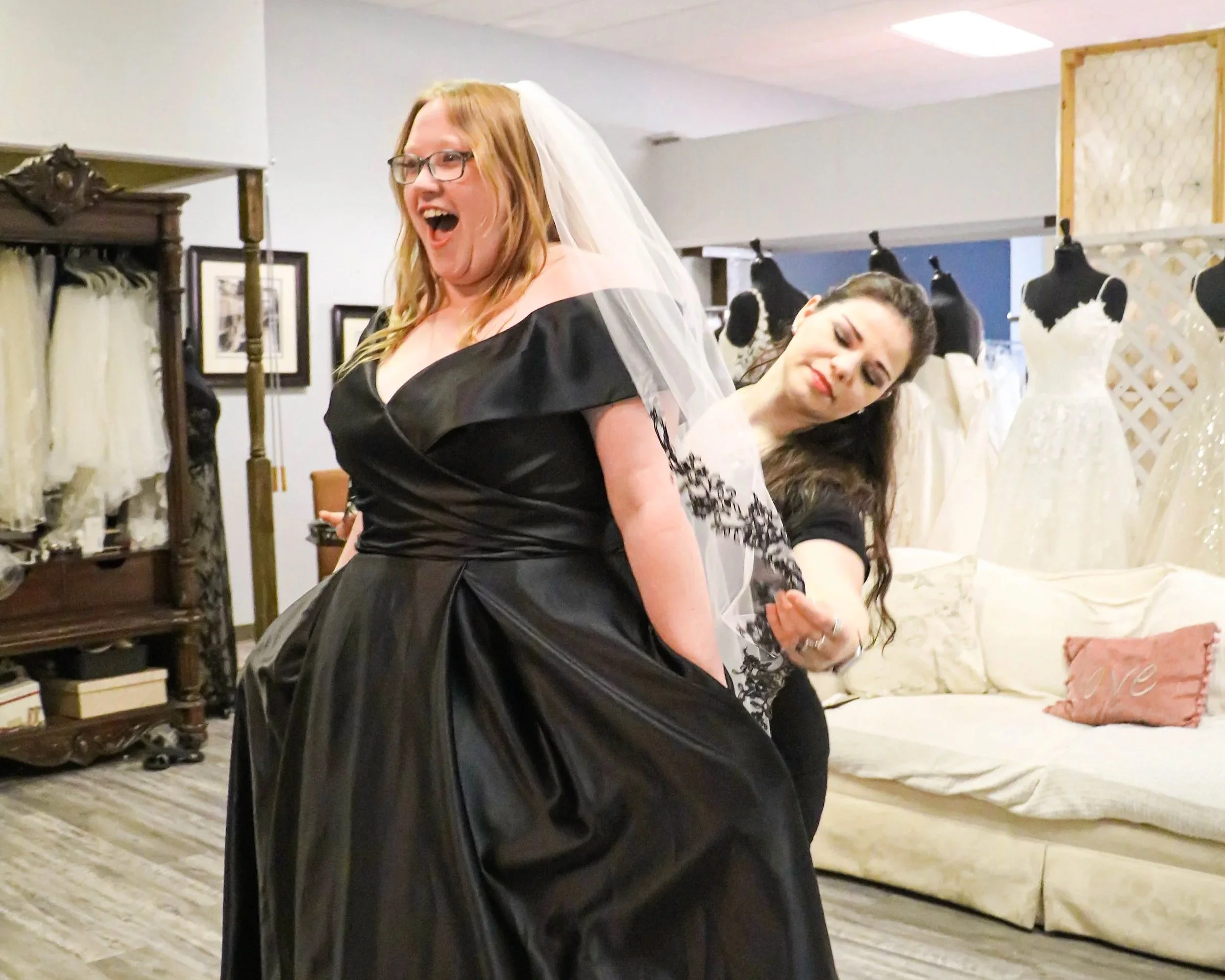 A bride-to-be smiles with excitement while trying on a black off-the-shoulder wedding gown and veil, as a bridal consultant makes adjustments during a fitting at a wedding dress boutique.