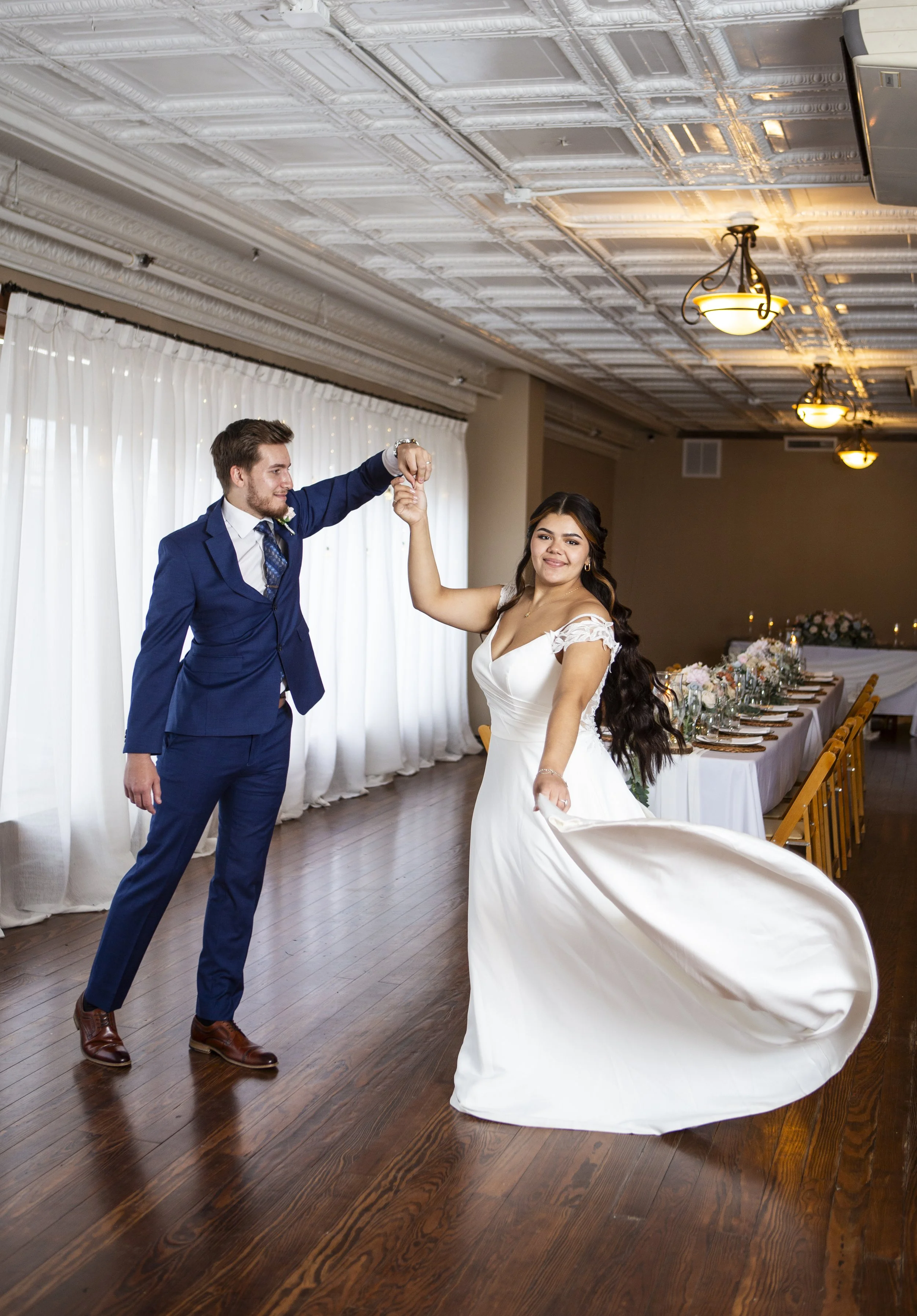 Bride in an elegant off-the-shoulder wedding gown spins across the dance floor as her groom in a blue suit leads the dance, capturing a romantic and joyful wedding reception moment.
