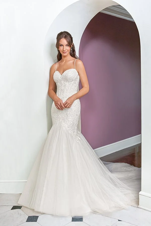 Bride models a lace mermaid wedding dress featuring a ruched sweetheart bodice, delicate spaghetti straps, and a flowing tulle skirt beneath a graceful archway, showcasing timeless elegance and romantic bridal fashion.