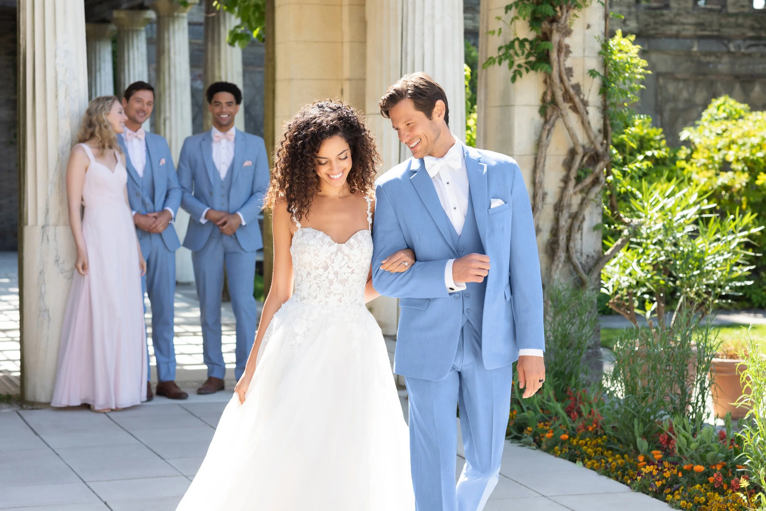 Bride in a romantic lace wedding gown strolls with her groom in a light blue three-piece suit through an elegant garden venue, with coordinated wedding party members and classical architecture creating a timeless wedding scene.