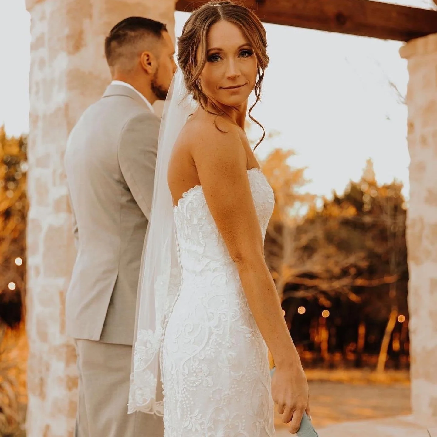 Bride wearing a strapless lace wedding gown and veil poses during a golden hour wedding portrait, with her partner in a light suit standing nearby at an elegant outdoor ceremony location.