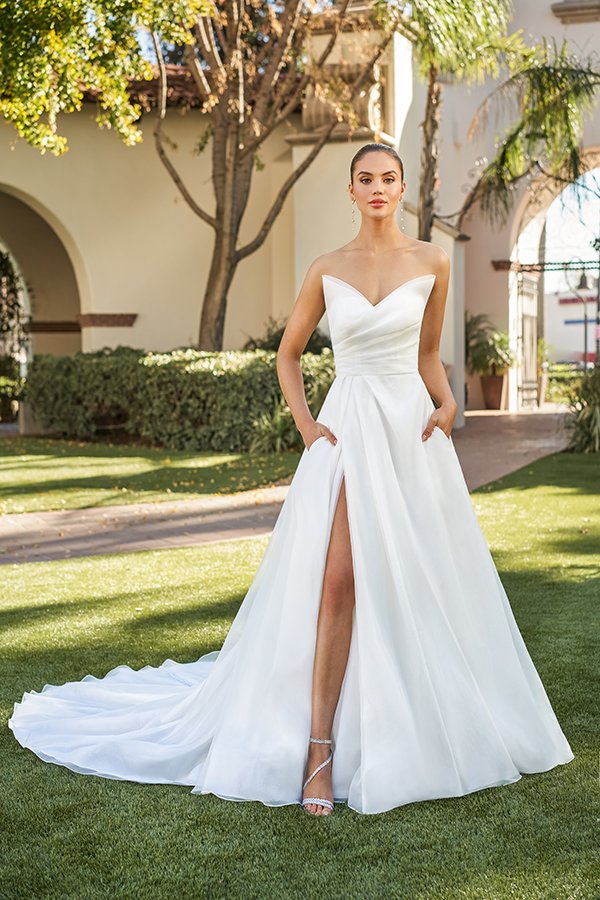 Bride modeling a modern strapless A-line wedding dress with a sweetheart neckline, softly draped bodice, hidden pockets, a thigh-high slit, and a flowing chapel train, photographed in a beautiful outdoor garden courtyard.