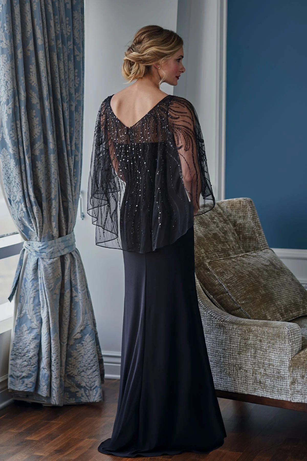 Back view of a black mother of the bride gown with a sparkling beaded illusion cape overlay, flowing sheer sleeves, V-back detail, and a sleek floor-length silhouette for formal occasions.