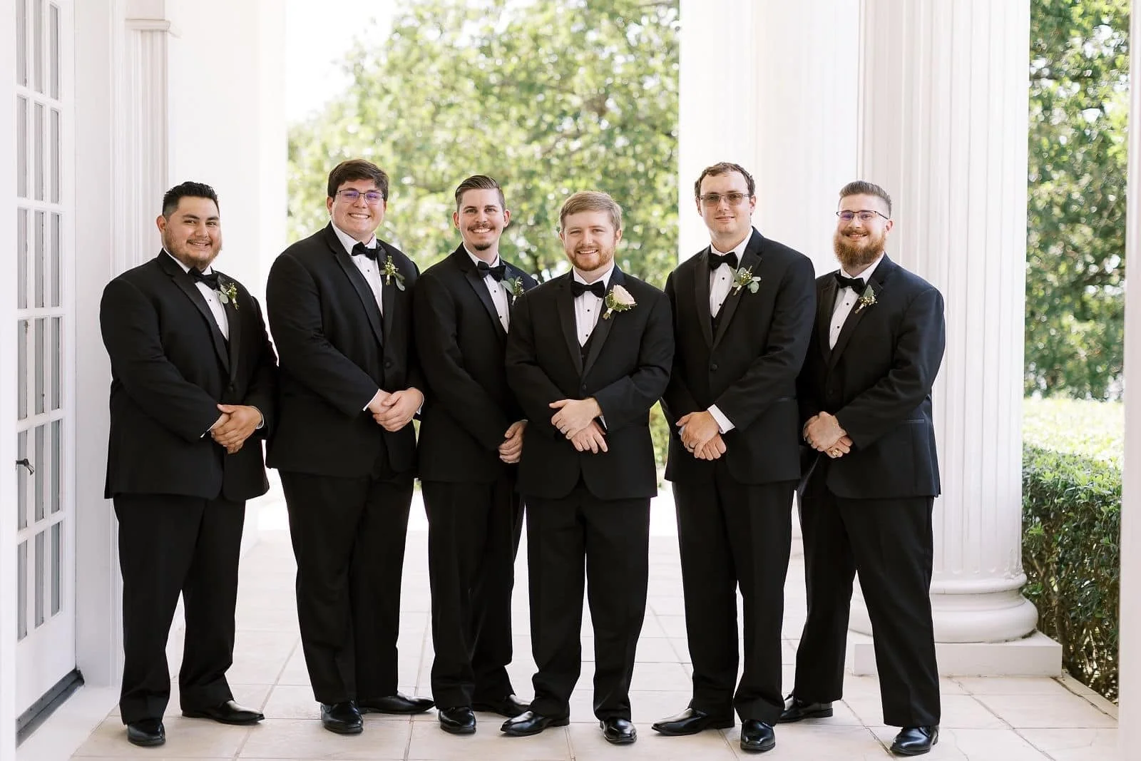 Groom and his groomsmen in coordinated black tuxedos and bow ties pose beneath grand white columns, creating a timeless and sophisticated wedding party portrait at a refined outdoor venue.
