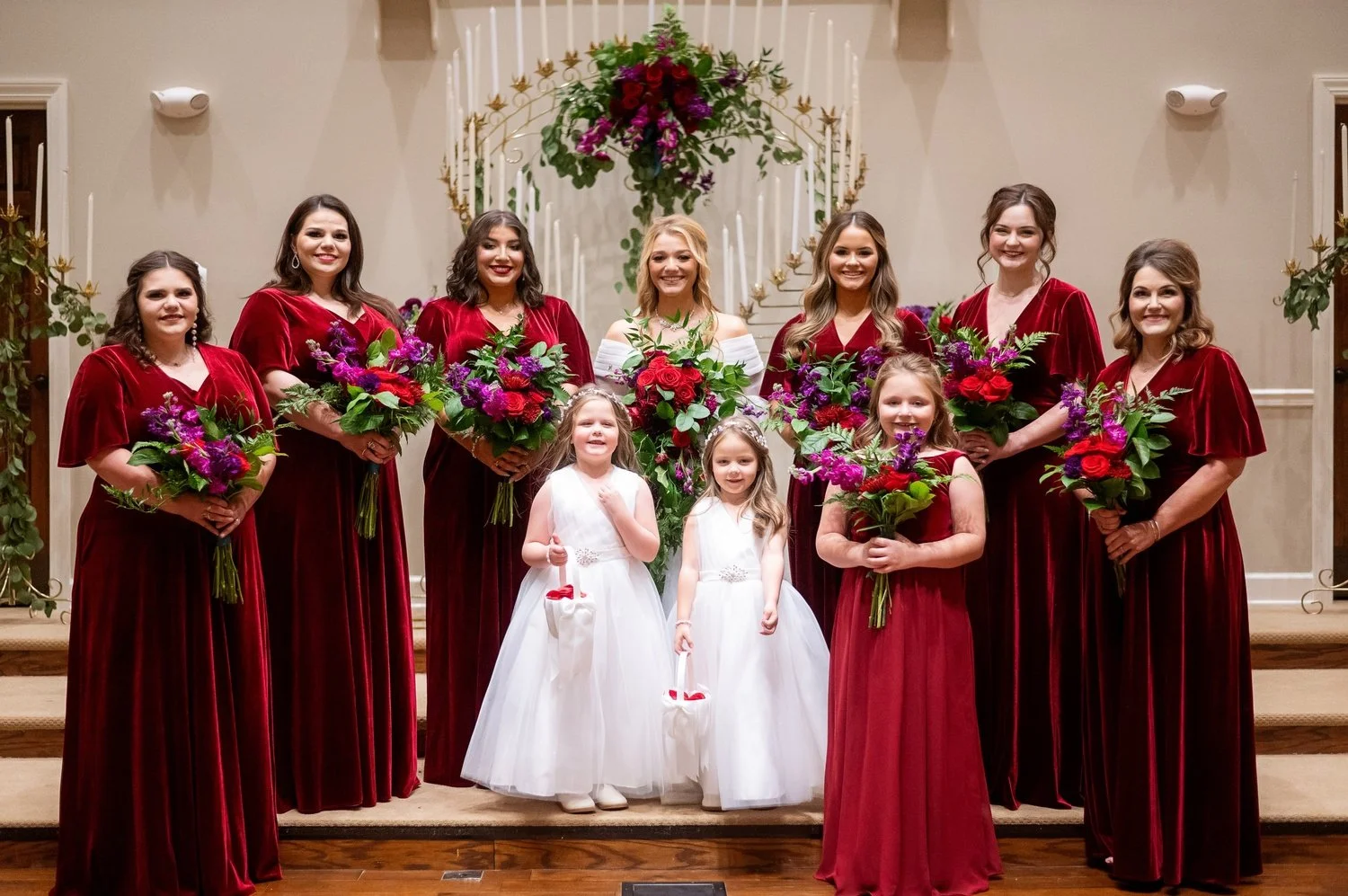 Bride poses with her bridal party featuring bridesmaids in elegant burgundy velvet gowns and flower girls in white dresses, holding bold red, pink, and purple bouquets in a beautifully decorated ceremony setting.