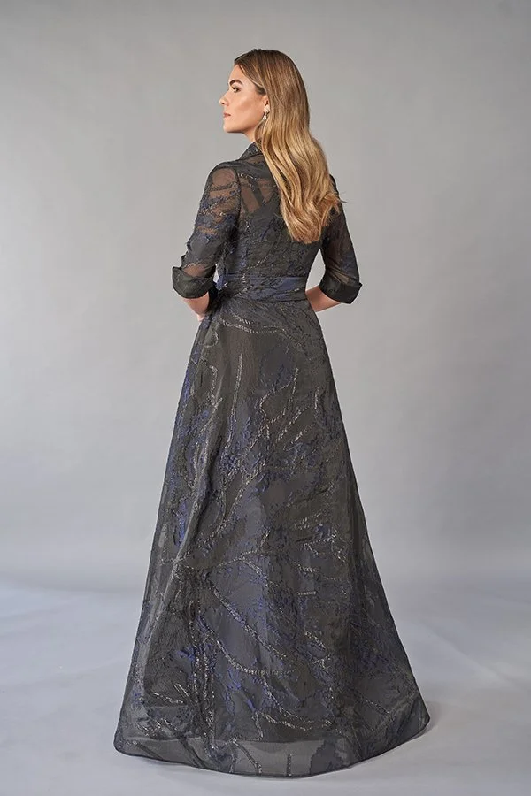Back view of an elegant charcoal mother of the bride gown with a collared neckline, sheer three-quarter sleeves, metallic cobalt jacquard fabric, waist tie belt, and a flowing A-line silhouette for formal occasions.