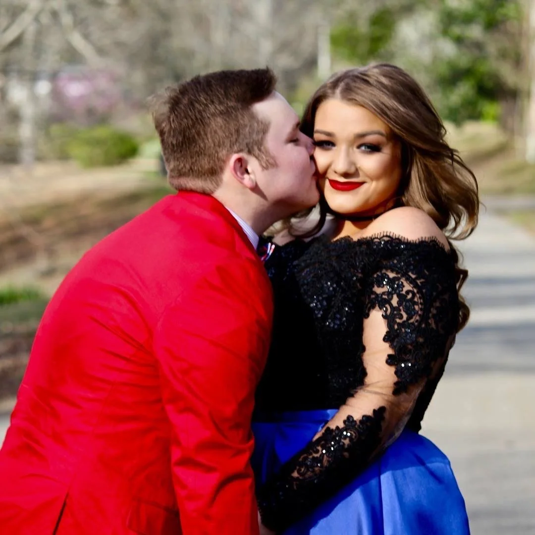 A young couple shares a sweet prom moment outdoors as the young man in a red formal jacket kisses his date’s cheek. Her elegant black lace off-the-shoulder gown with a vibrant blue accent creates a striking and romantic formal portrait.